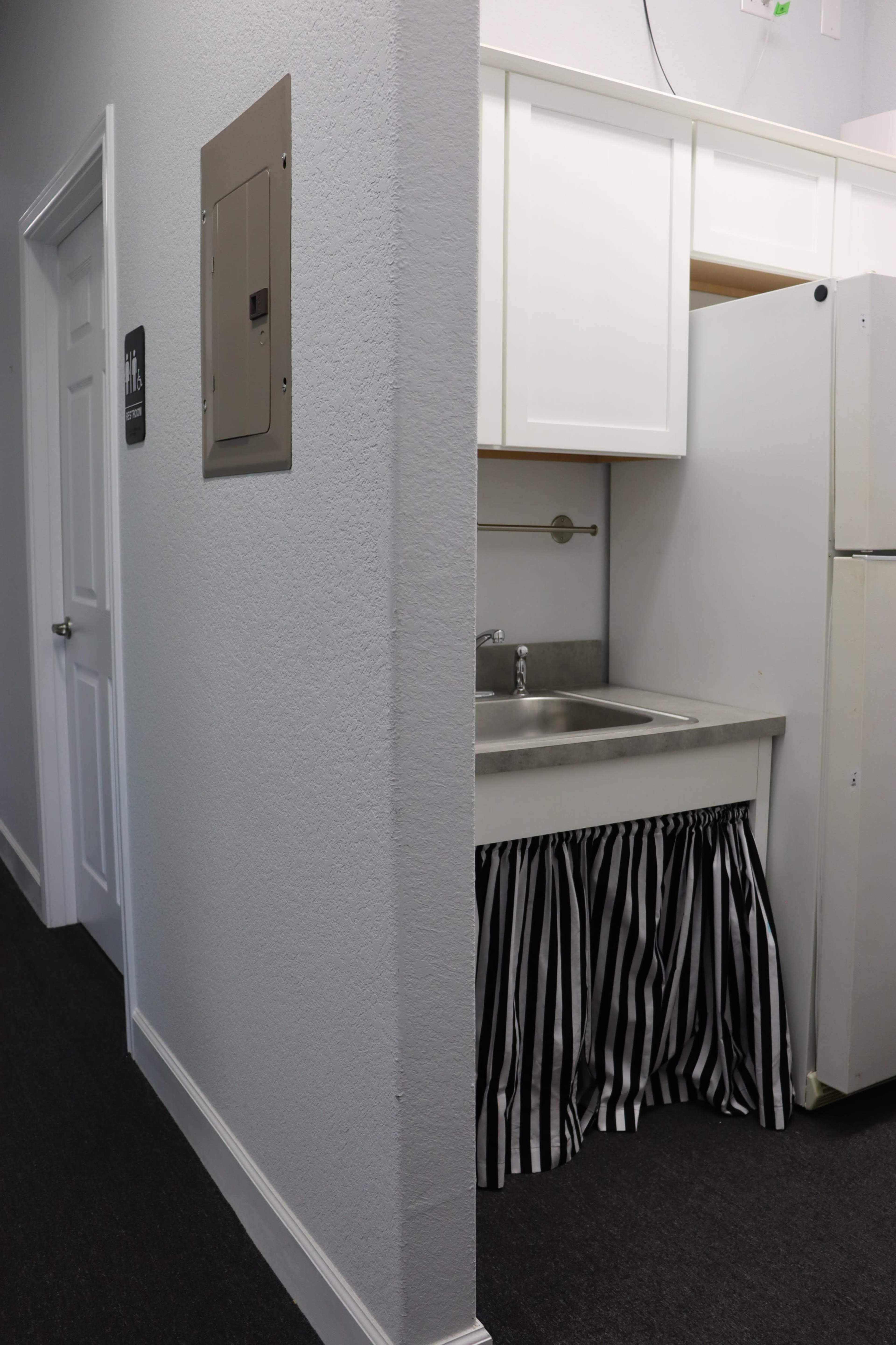 A small kitchenette with a sink and a white refrigerator, partially visible through an opening in a wall beside a door labeled for restrooms.