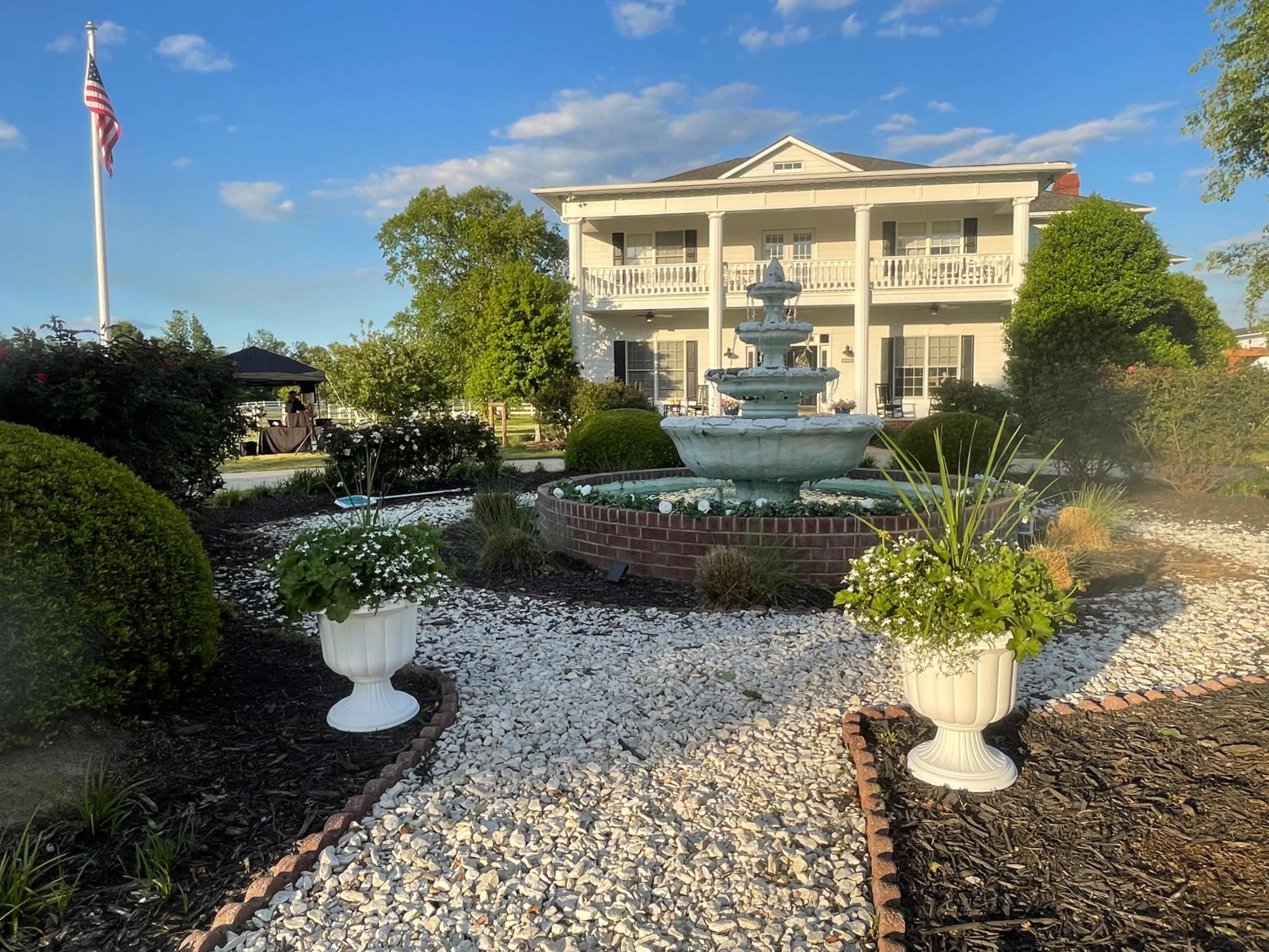 A multi-tiered fountain is surrounded by a landscaped garden featuring potted plants and a large colonial-style house in the background.