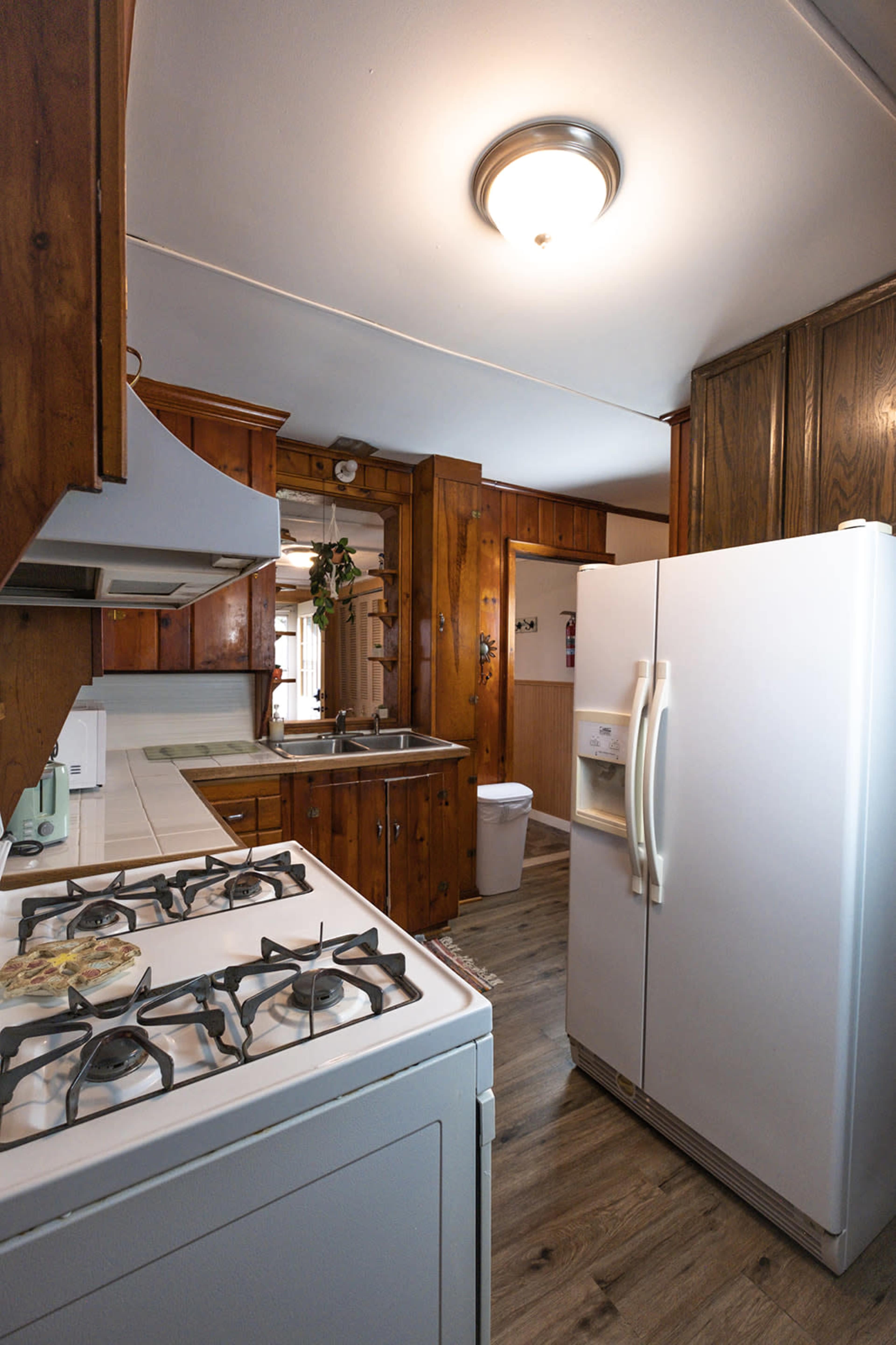 The image shows a kitchen with wooden cabinets, a white refrigerator, a gas stove, and a sink, all arranged in a compact space.