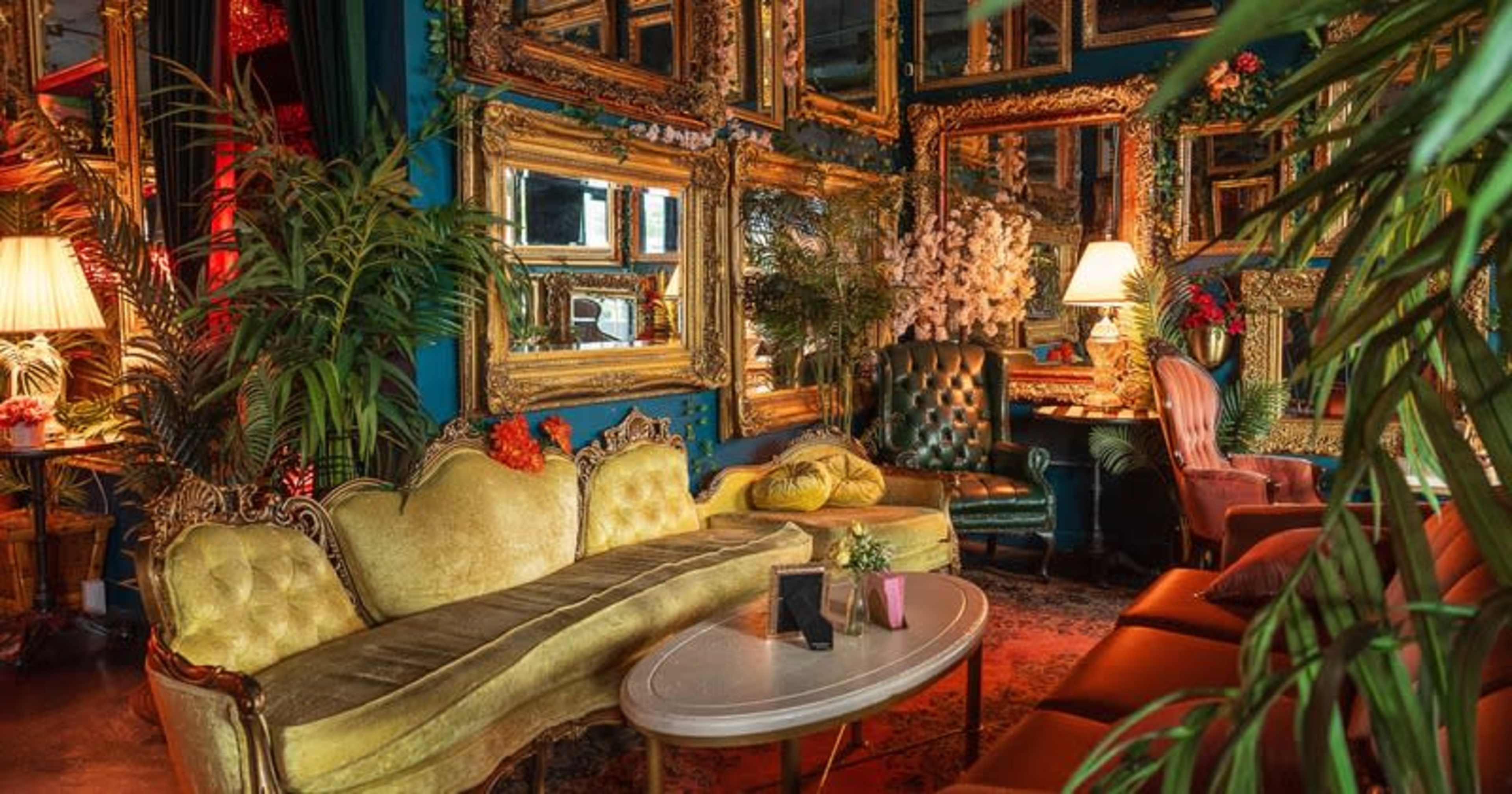 The image shows a vintage-themed lounge featuring ornate mirrors, plush seating, and various indoor plants.