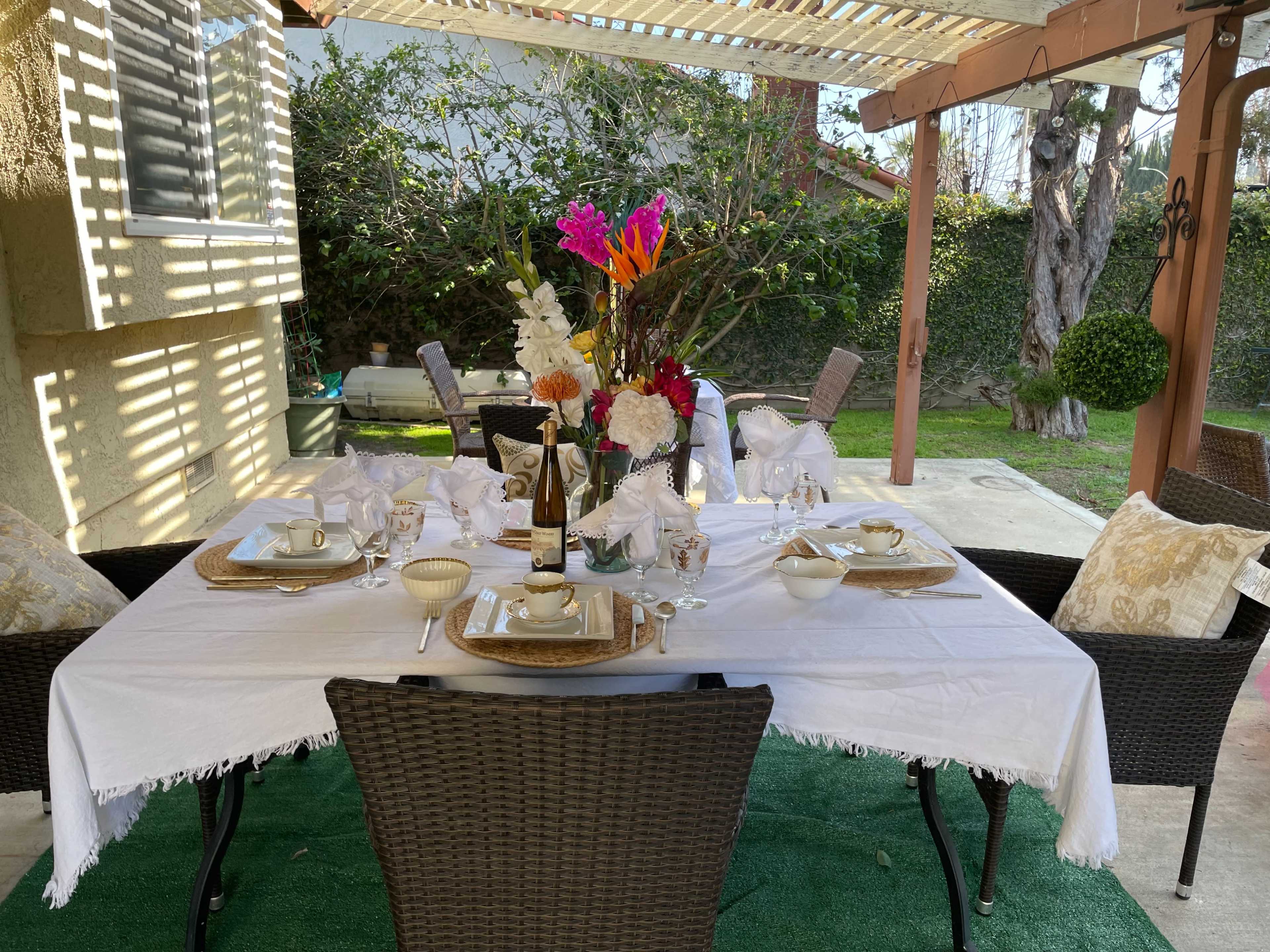 A nicely arranged outdoor dining table is set under a pergola, featuring a white tablecloth, elegant dishware, and vibrant floral centerpieces.