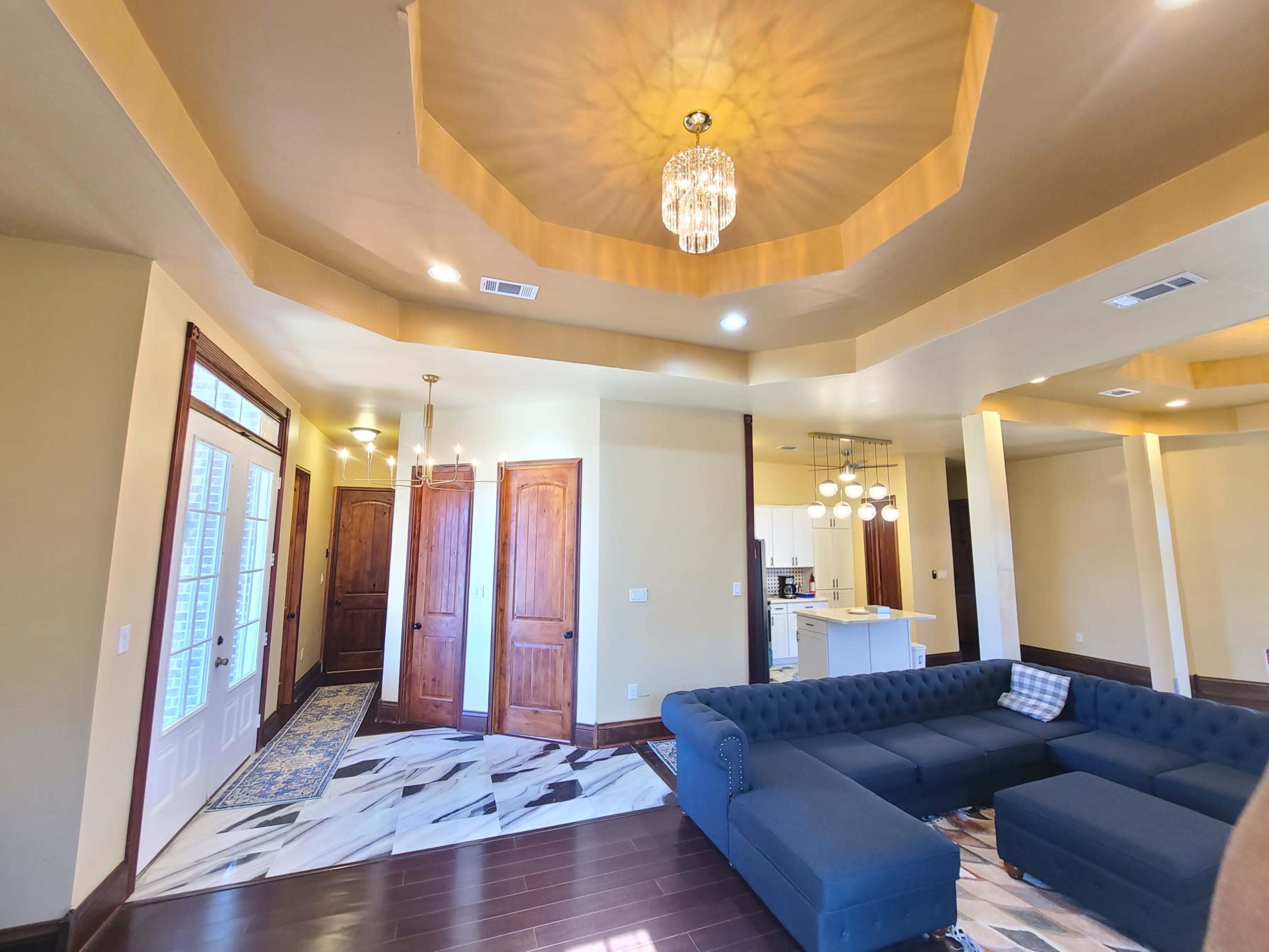 The room features a spacious living area with a blue sectional sofa, hardwood floors, and an entrance leading to a kitchen area.