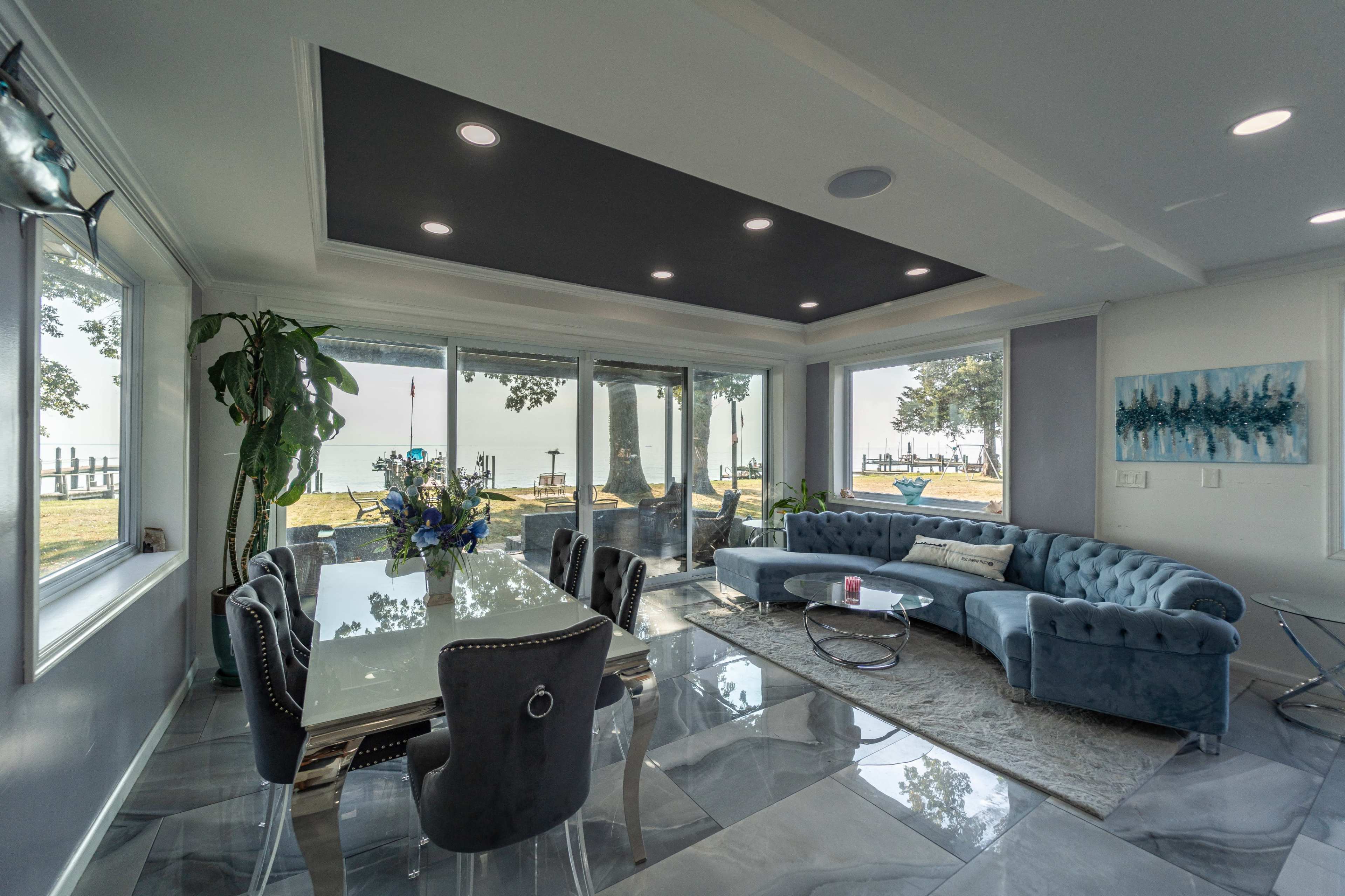 A modern living room features a glass dining table, a blue sectional sofa, and large windows overlooking a waterfront view.