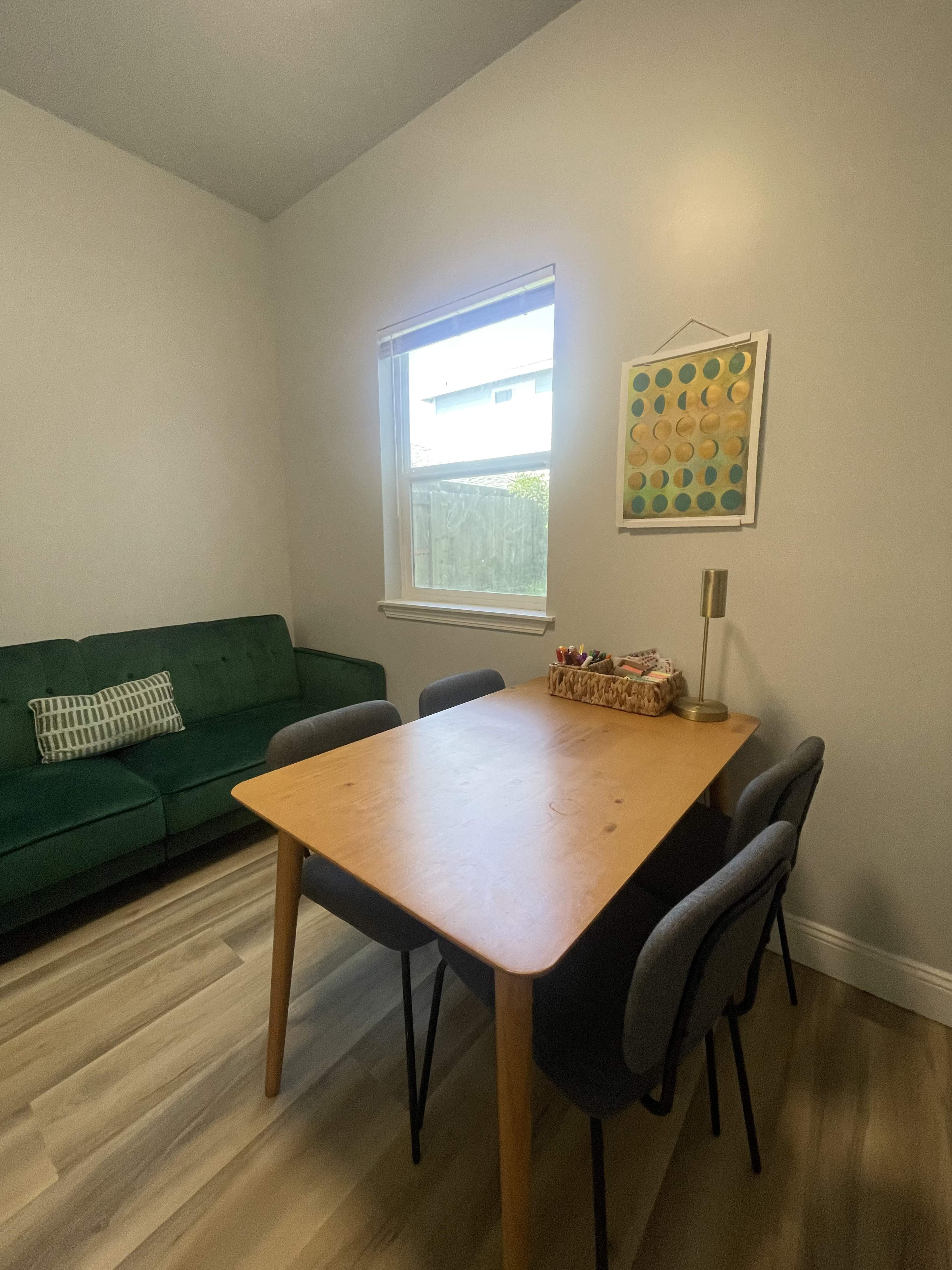 A small dining area with a wooden table surrounded by four gray chairs, a green couch is positioned against the wall, and a window lets in natural light.
