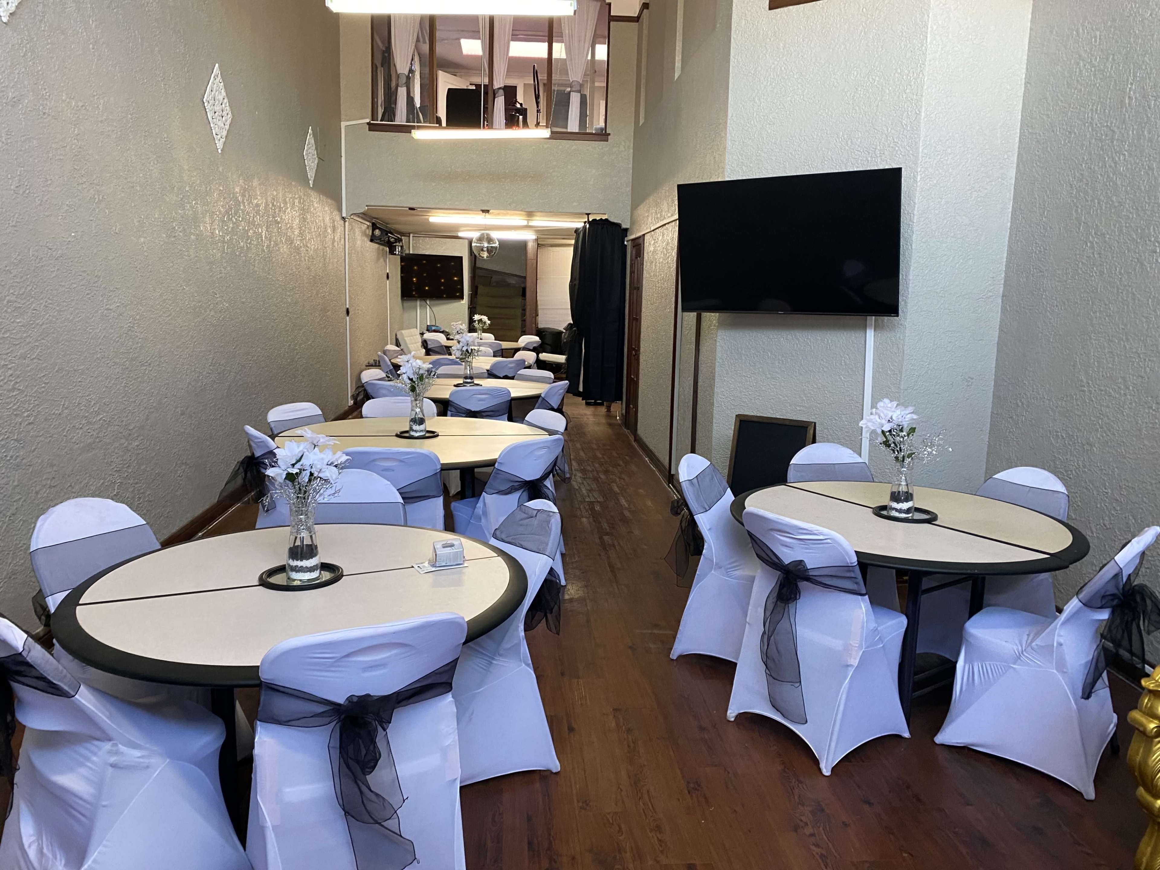 A decorated event space with round tables covered in white tablecloths and black sashes, arranged neatly along the length of the room.