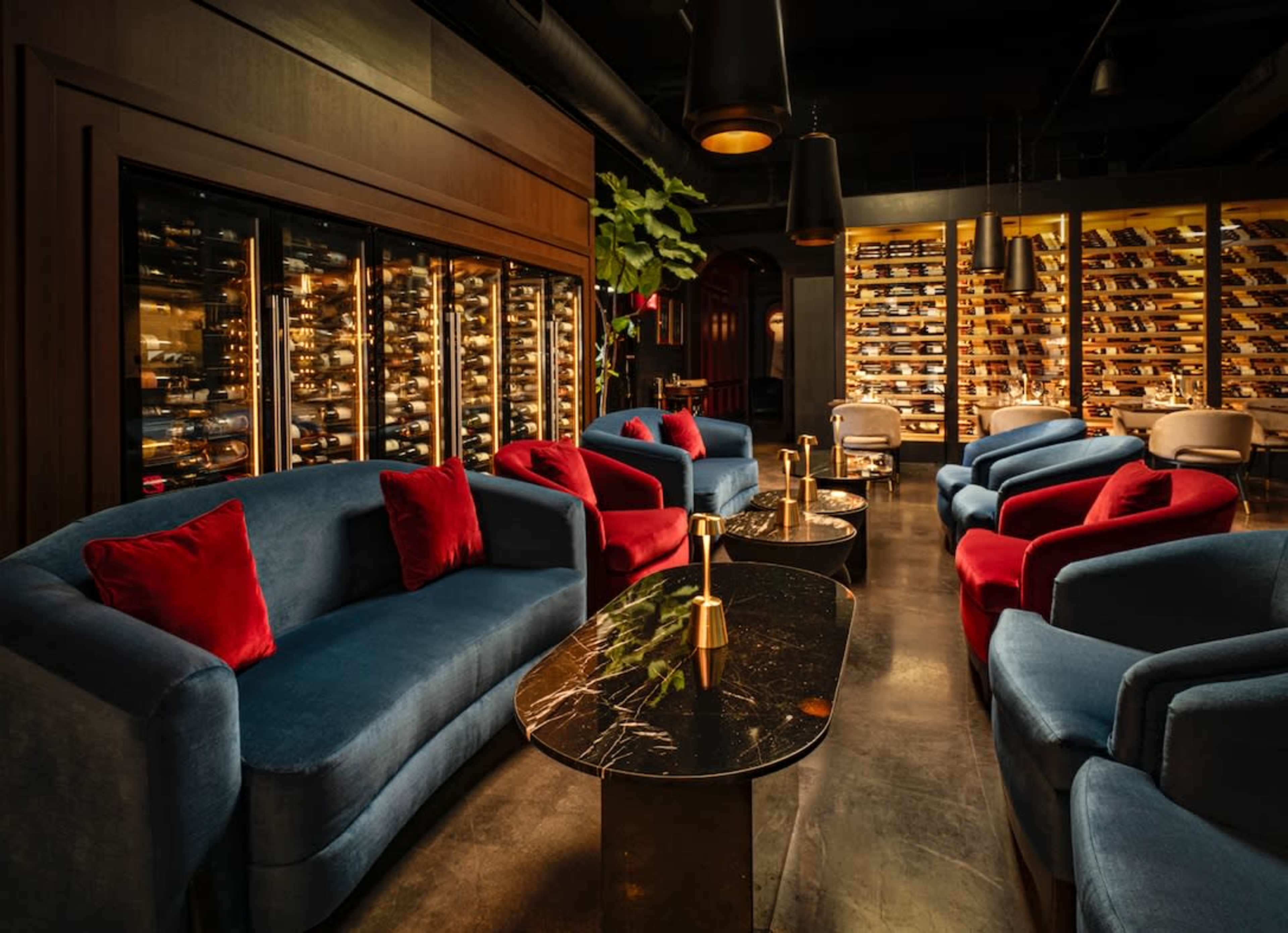 The image depicts a stylish lounge area with blue and red upholstered sofas arranged around marble-topped tables and a wall of wine storage in the background.