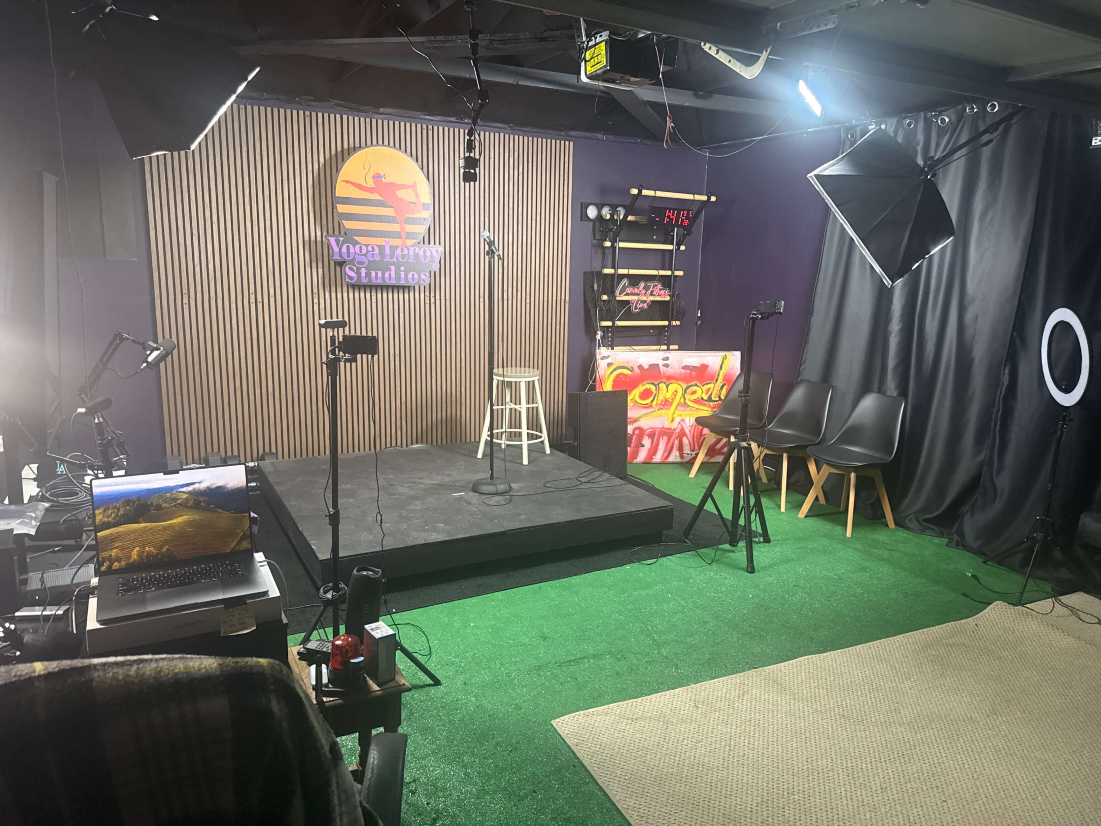The image shows a small indoor studio set up with a stage, microphone, lighting equipment, a laptop, and seating for an audience, featuring a green carpet background.