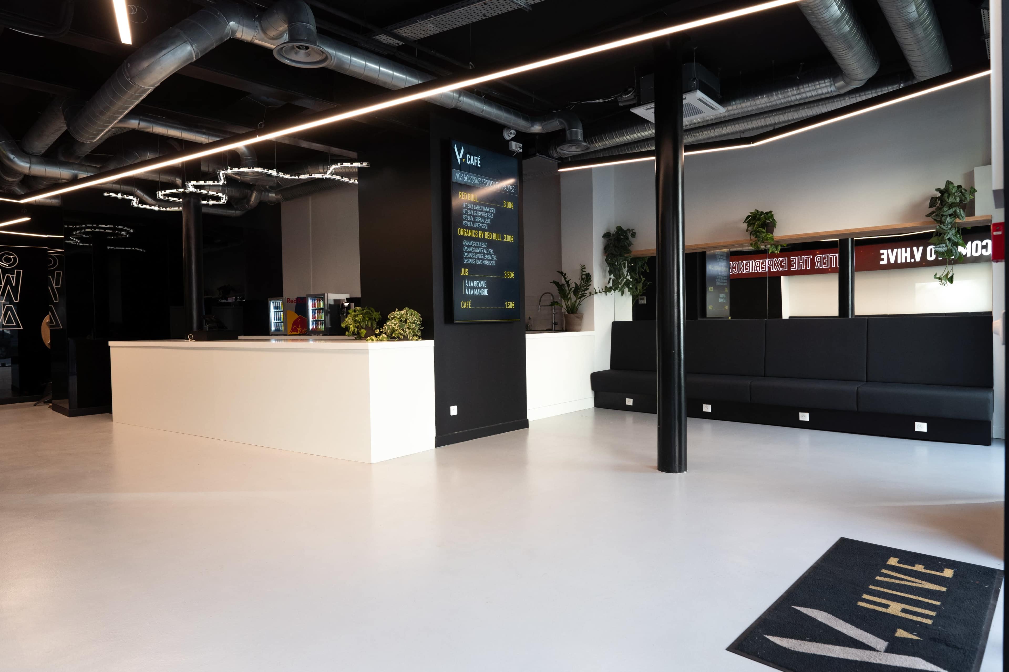 The image shows a modern reception area with a white counter, black seating along the walls, and a large screen displaying information.