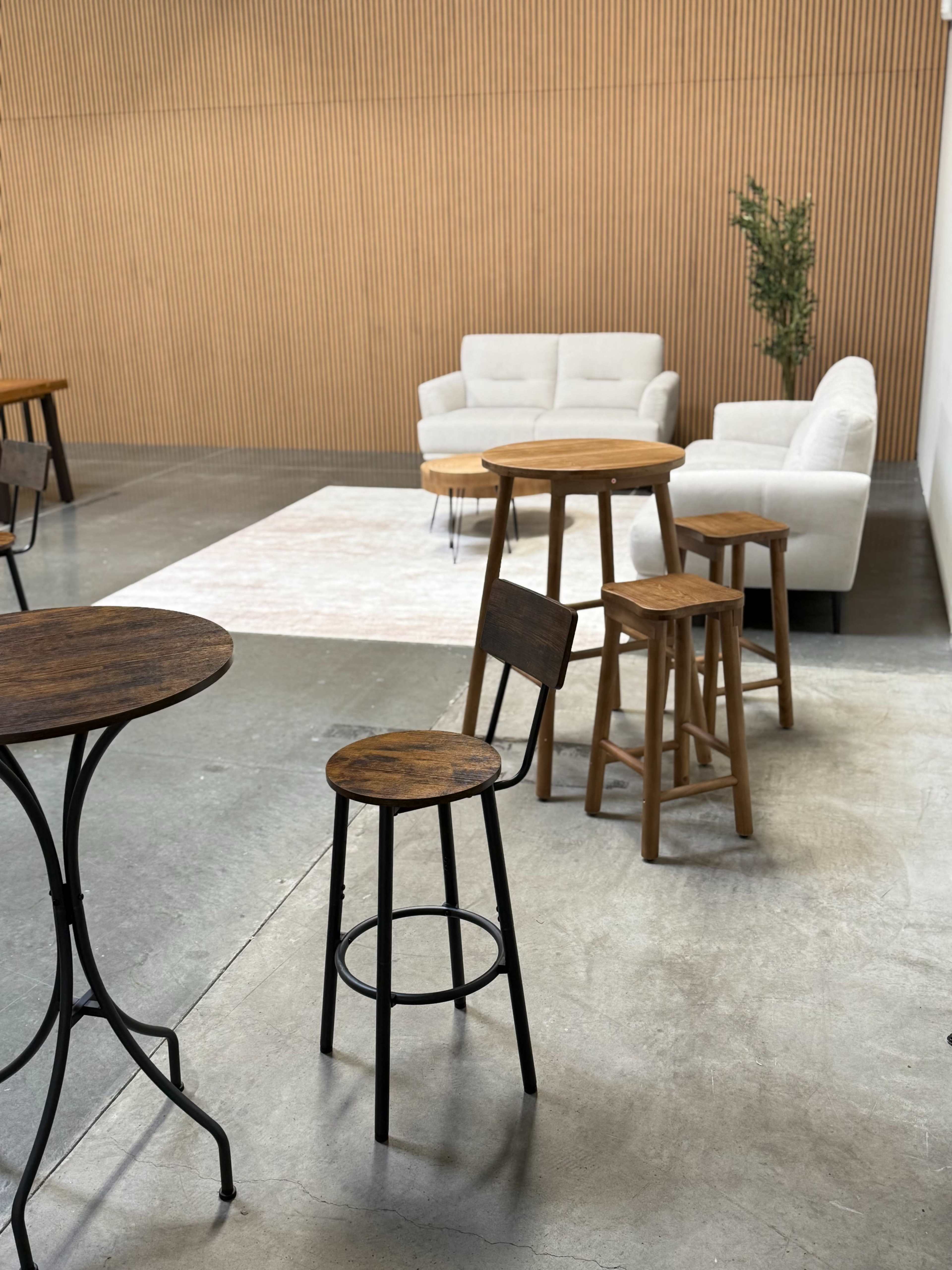 The image shows a modern interior with wooden stools and tables arranged on a concrete floor, alongside a white sofa and a decorative plant against a wooden panel wall.