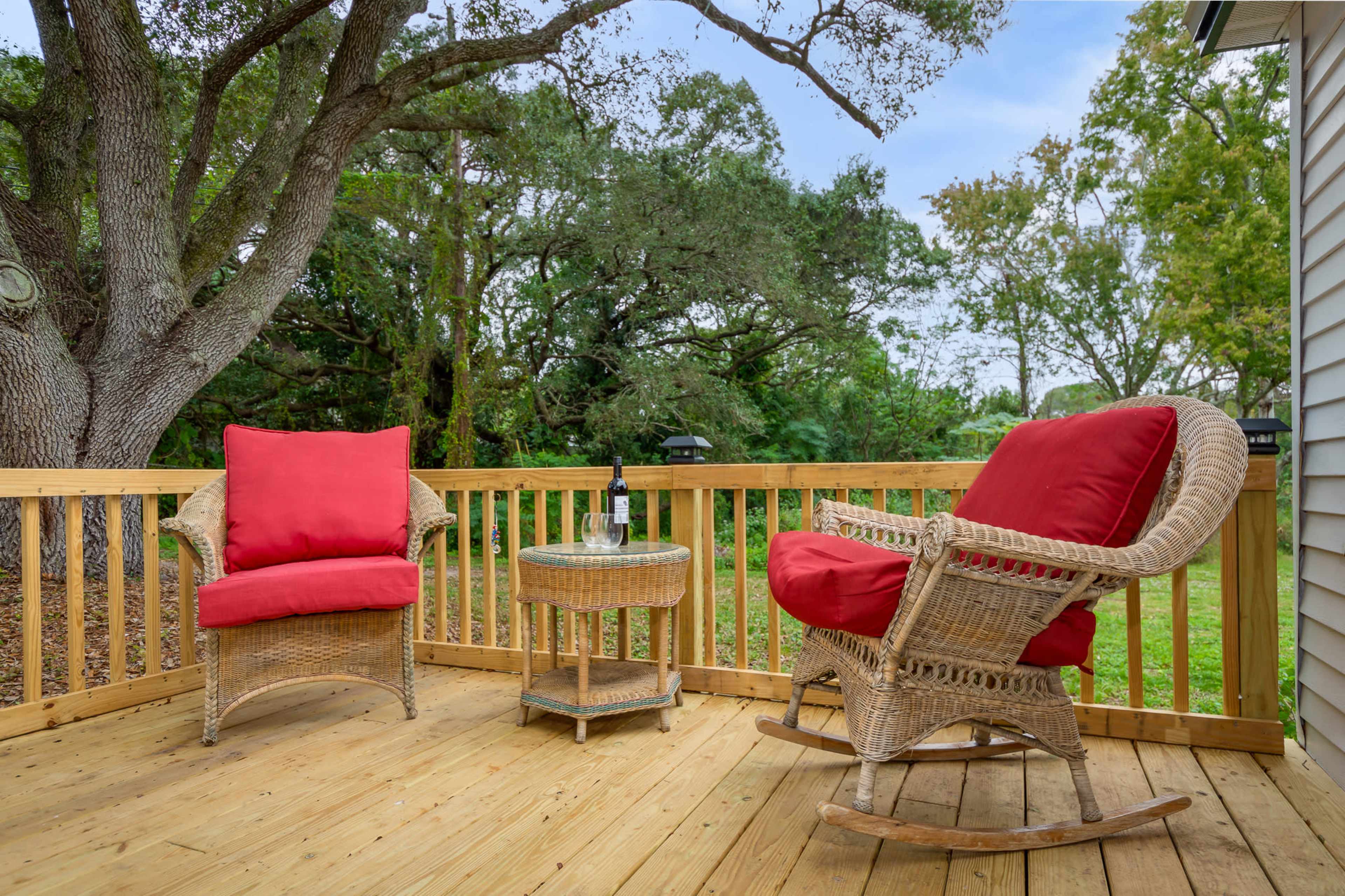 A wooden deck features two wicker armchairs with red cushions and a small round table beside a bottle of wine, surrounded by greenery.