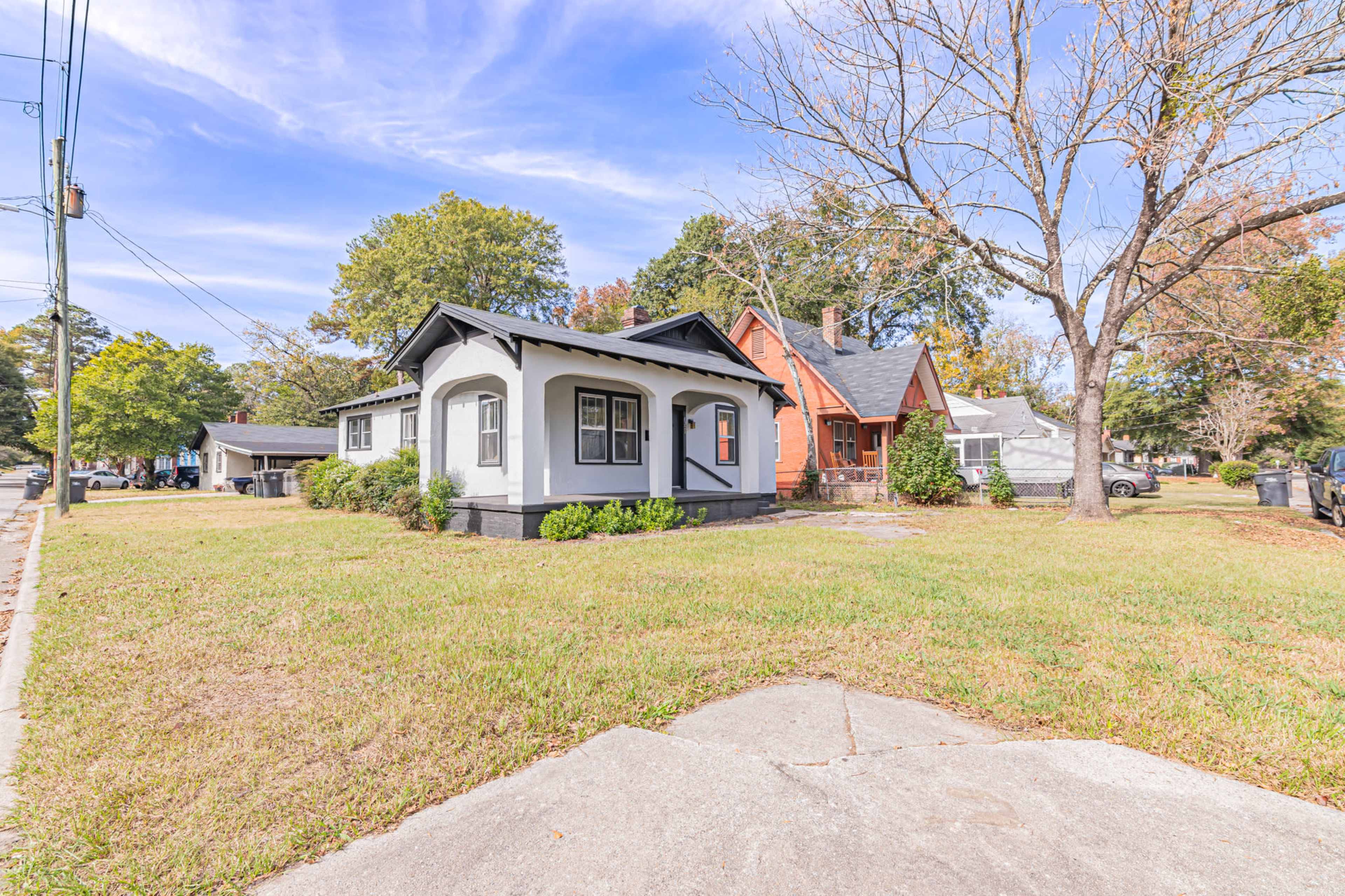 A street view featuring two houses, one with a gray exterior and a porch, and another with a red exterior, set on a grassy corner lot.