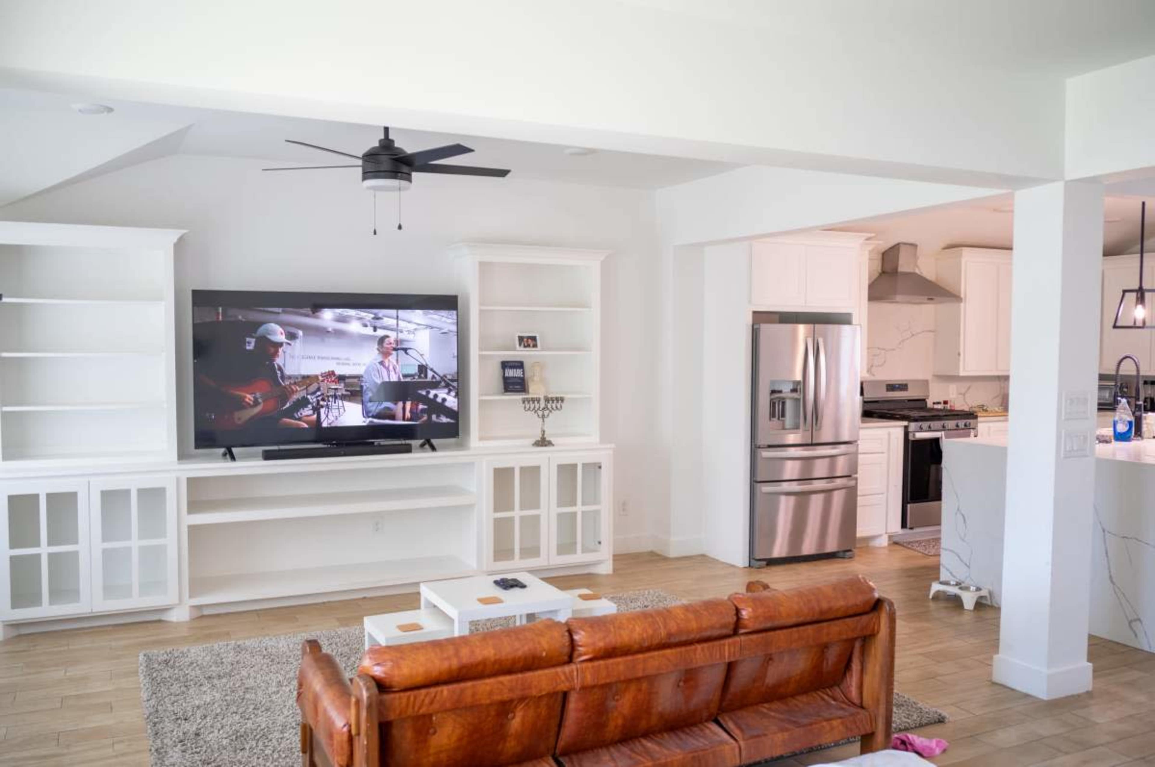 The image shows a modern living room with a large television displaying a performance, a brown leather couch, and an adjacent kitchen area featuring stainless steel appliances.
