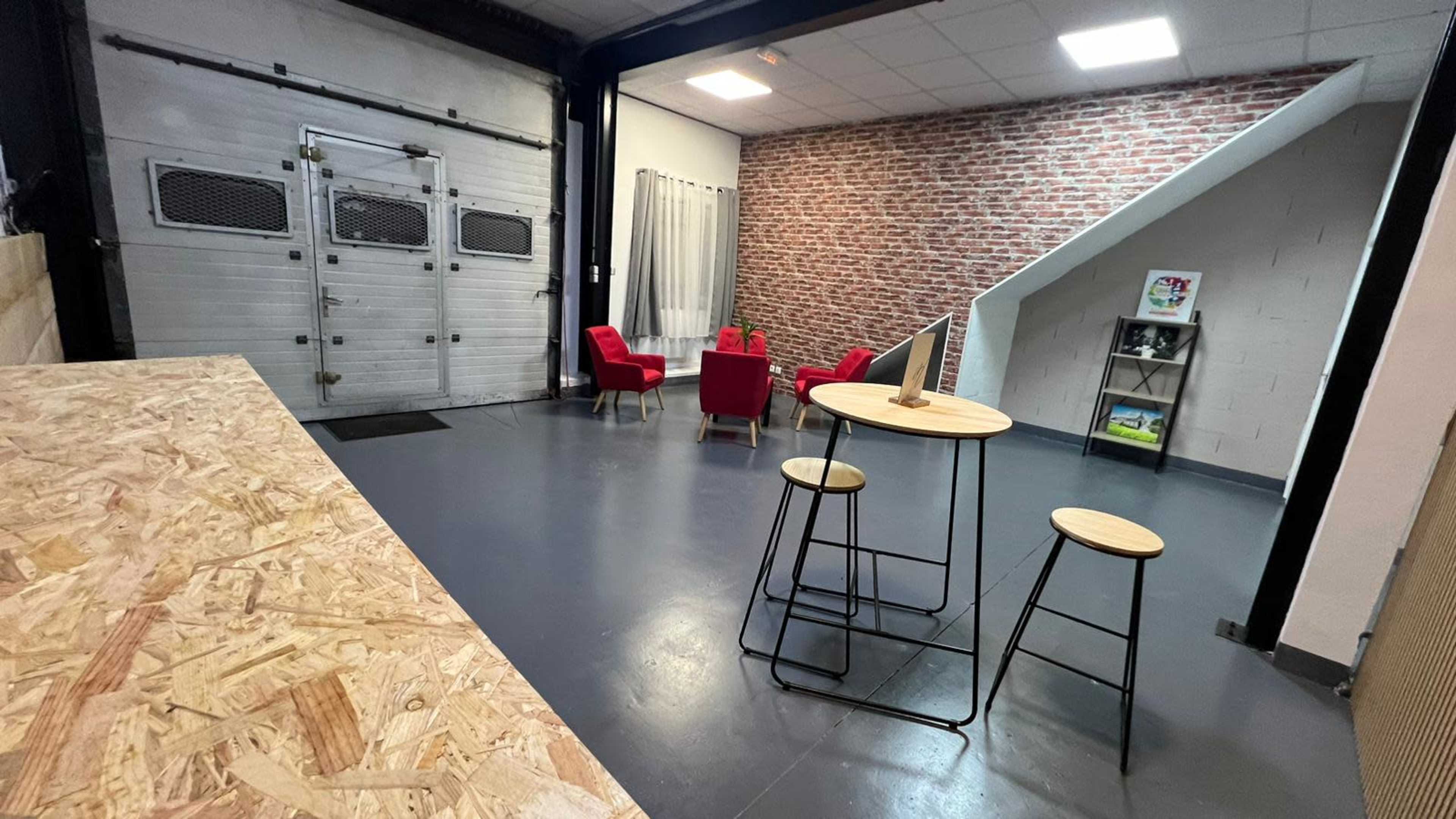 A modern indoor space featuring a wooden countertop, a table with three stools, a seating area with red chairs, and a brick-patterned wall.