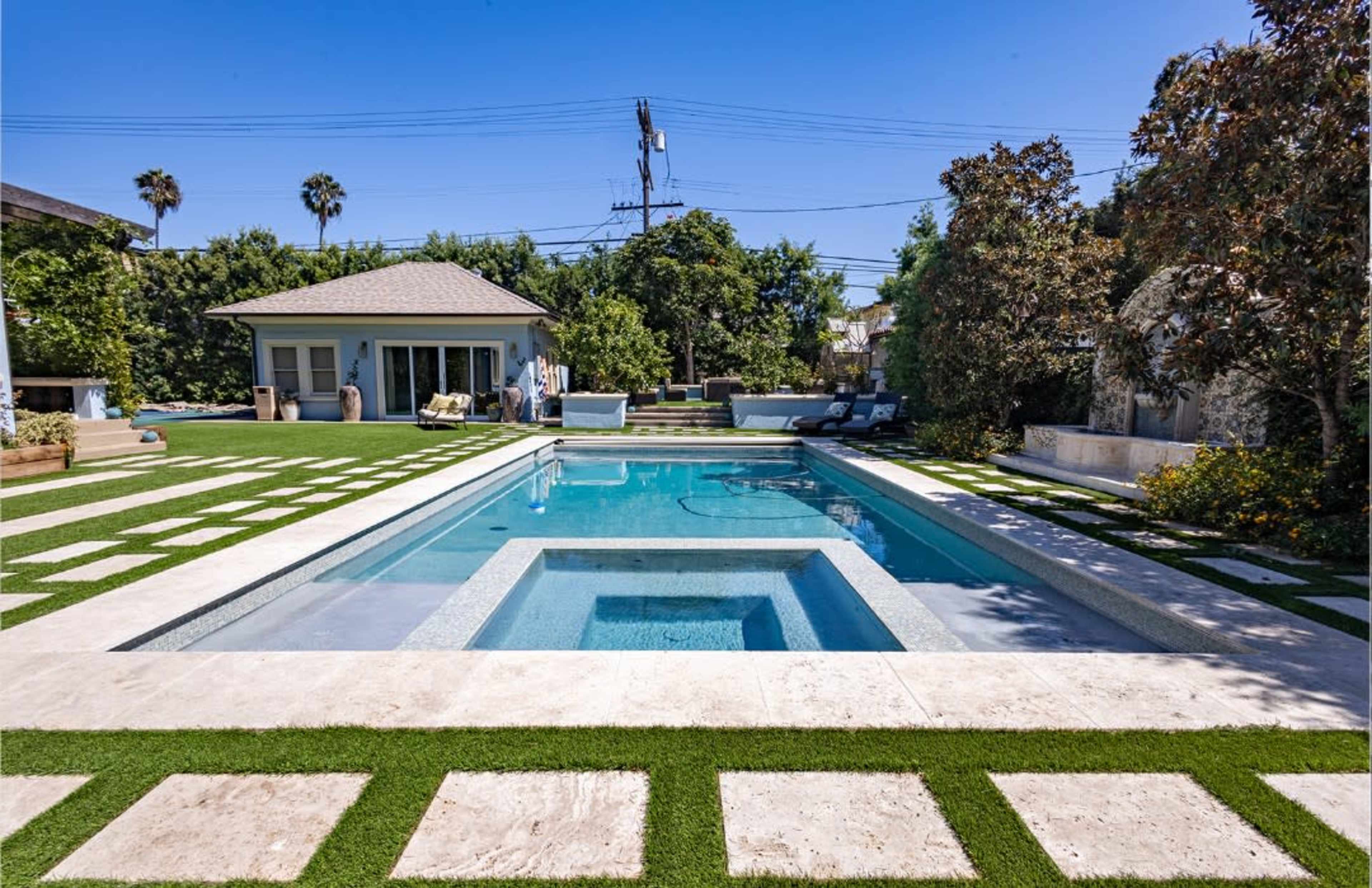 The image shows a modern backyard with a swimming pool, a hot tub, and a patio area surrounded by neatly arranged grass and decorative landscaping.