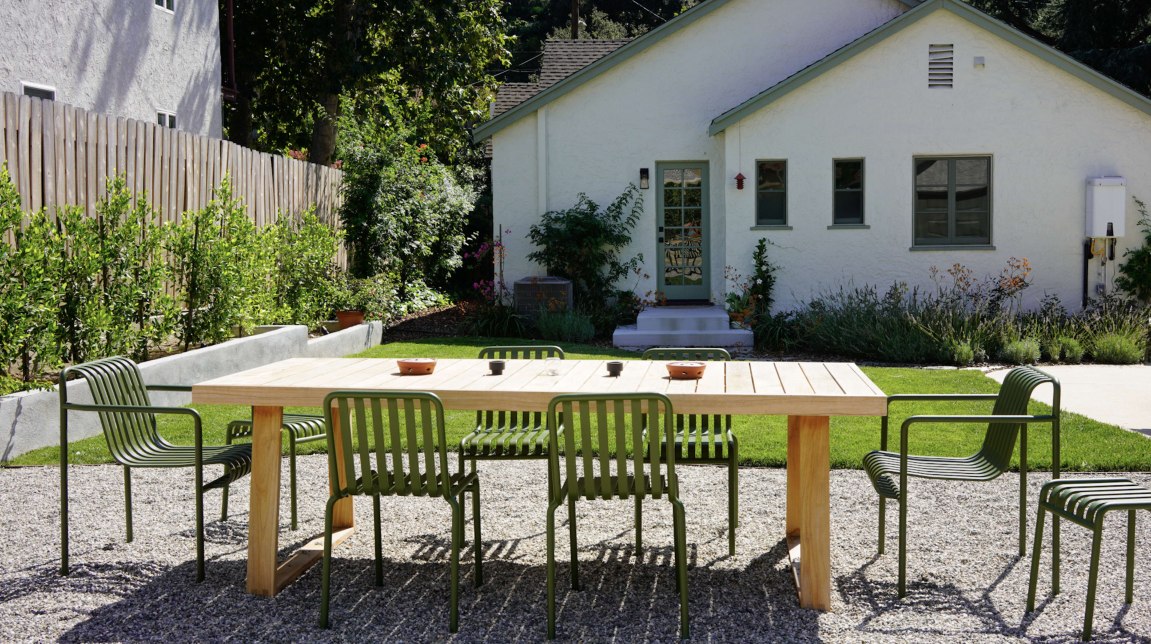 A wooden dining table with green chairs is set on gravel in a garden area, facing a white house with green window frames and a porch.