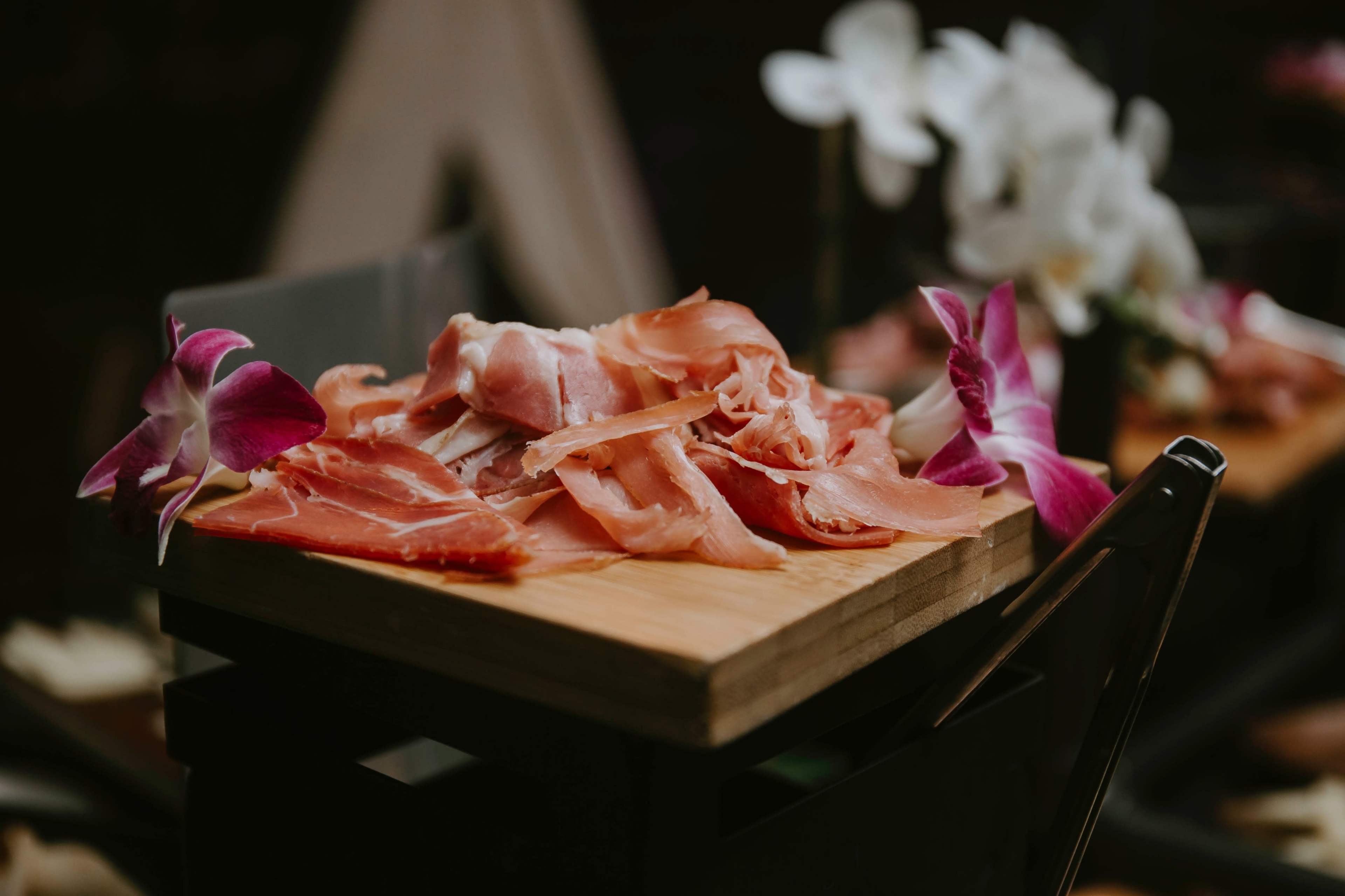 A wooden board displays various slices of cured meat surrounded by pink and white orchids.