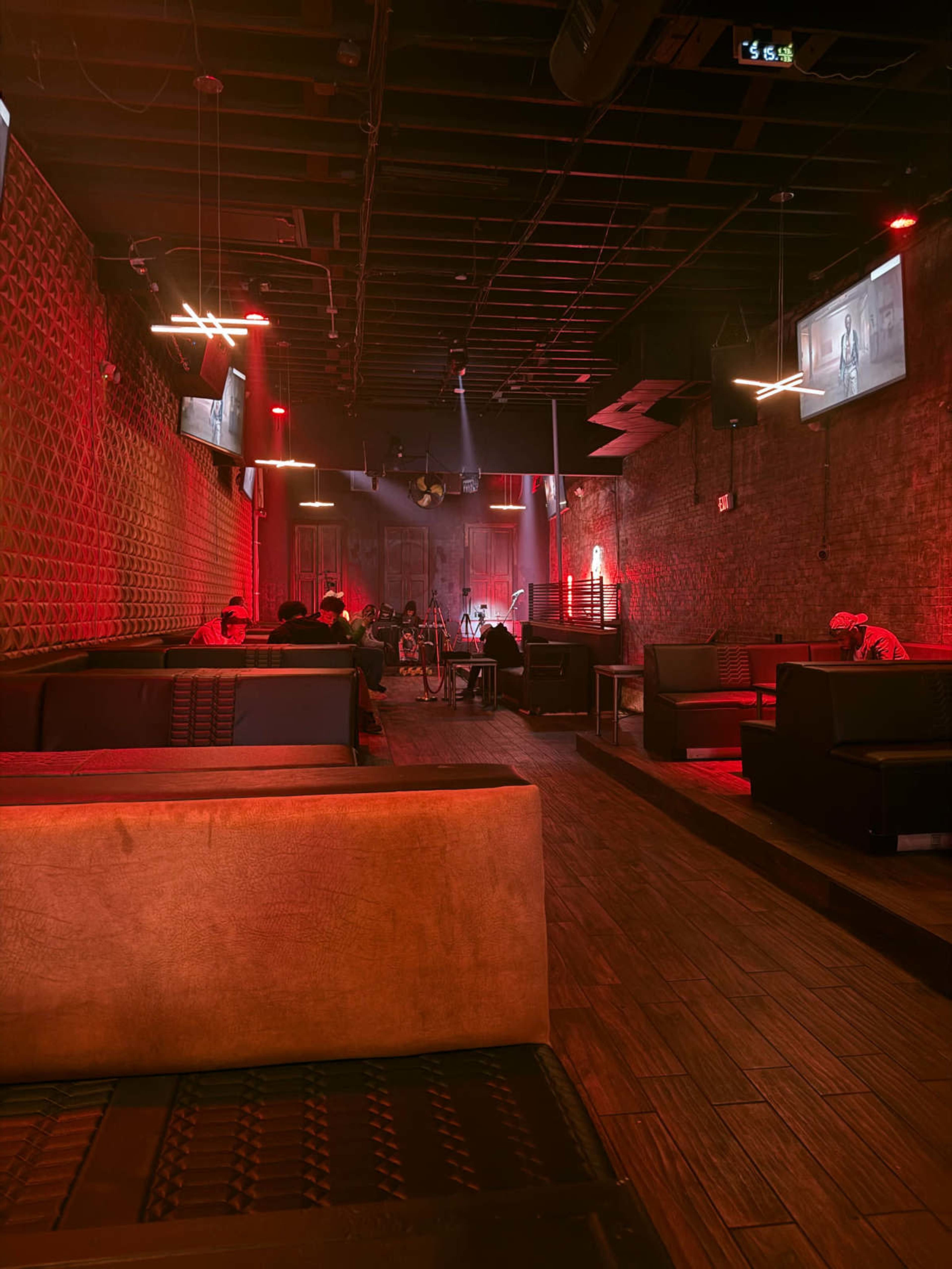 The image shows an empty lounge with red lighting, featuring dark furniture and a brick wall, alongside dimly lit tables and a stage area.