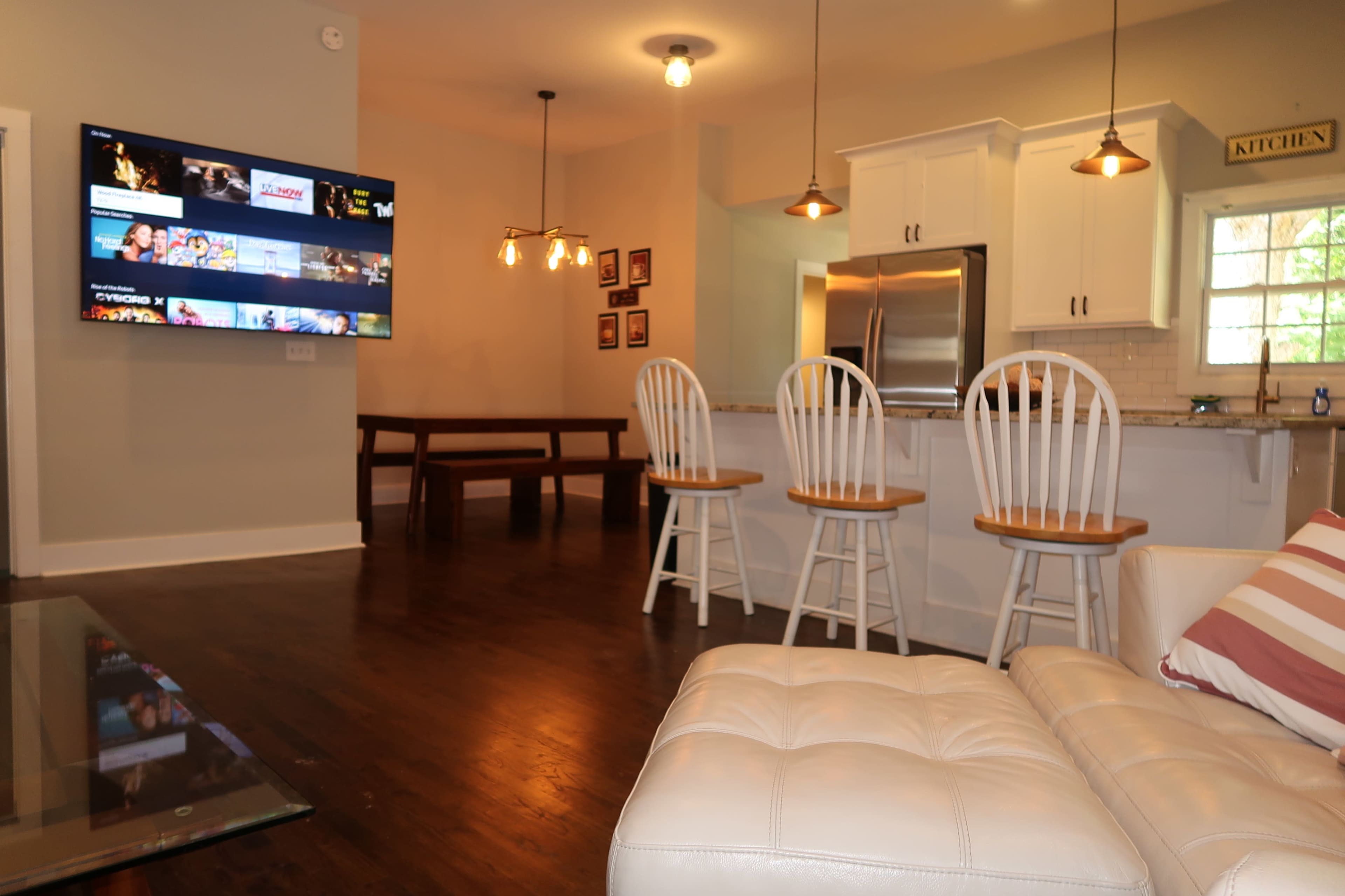 The image shows a modern living space featuring a kitchen area with white cabinetry, a dining table, and a seating area with a large flat-screen TV mounted on the wall.