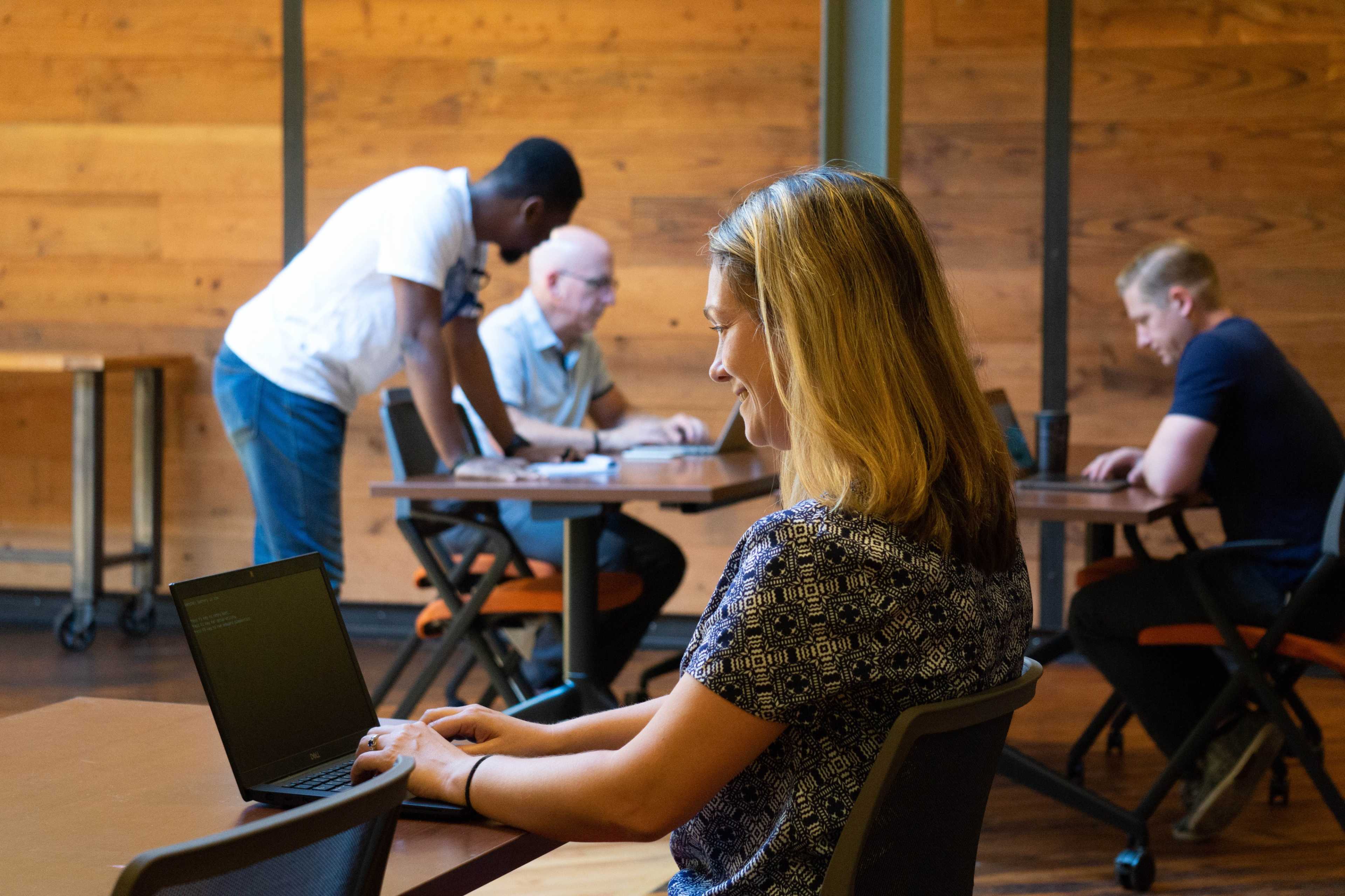 A woman works on a laptop while two men stand and sit at tables in a wooden-paneled room.