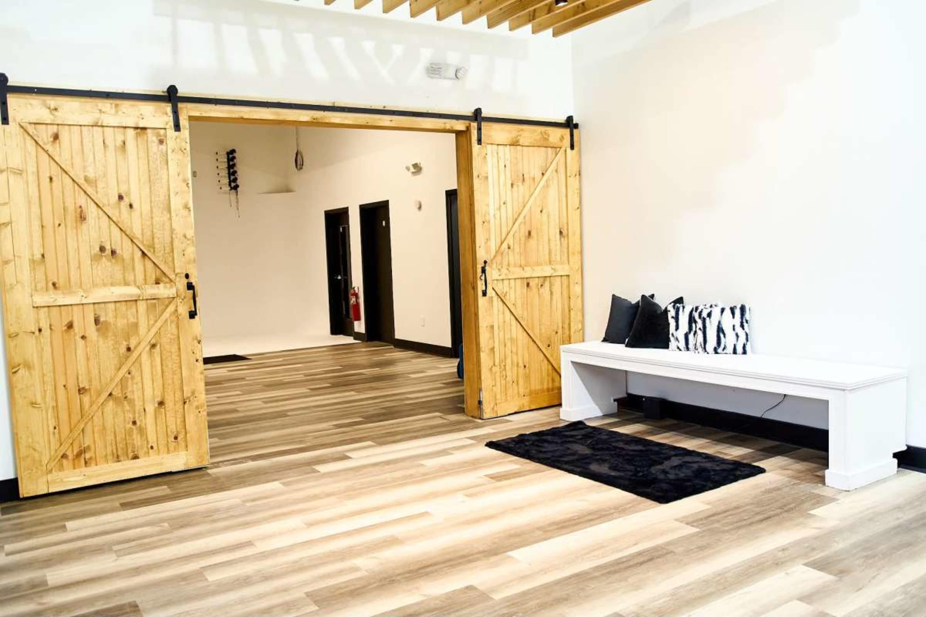A spacious room features large wooden sliding barn doors, a white bench, and a black rug on a floor of light-colored planks.