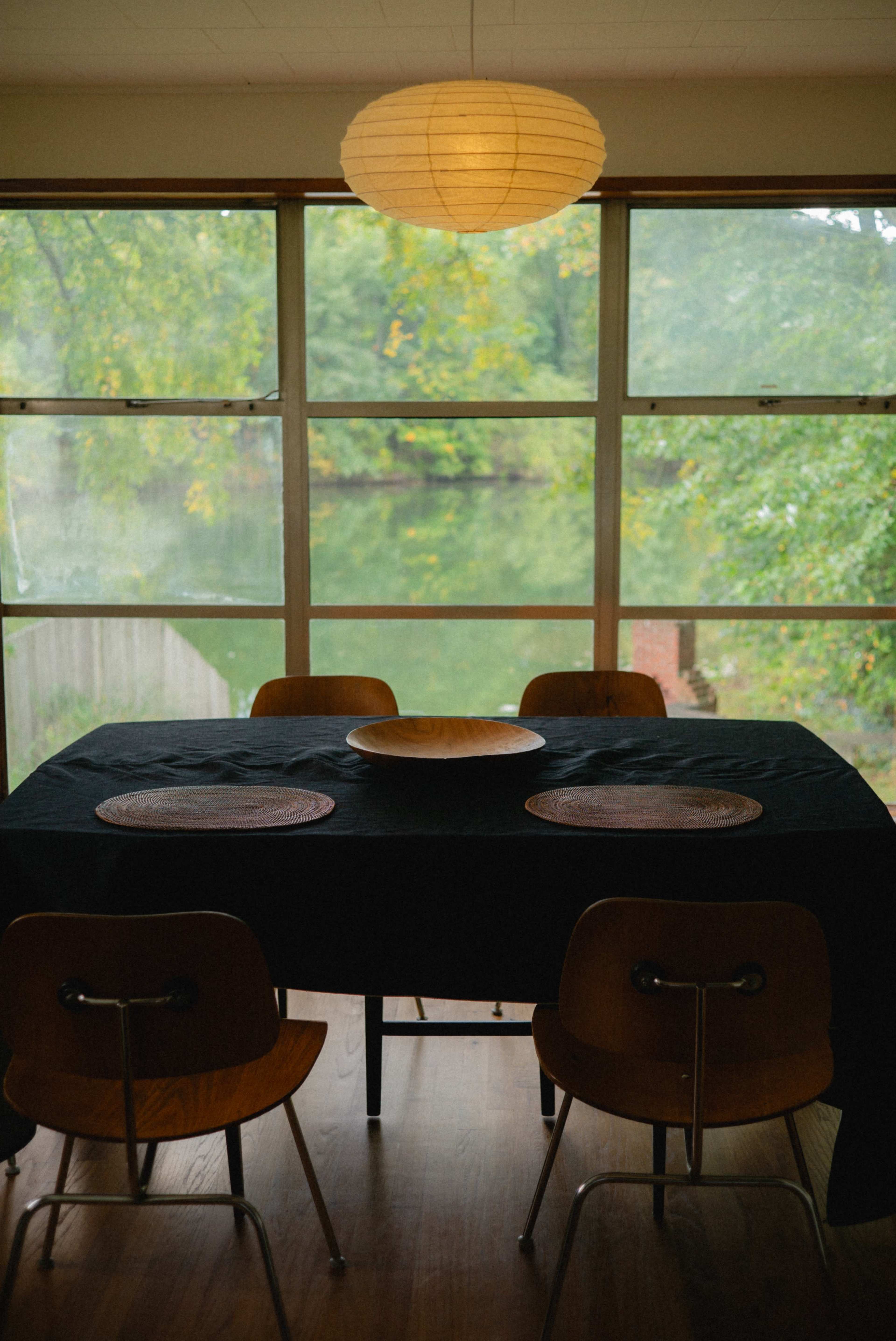 A wooden dining table with a black tablecloth is set with woven placemats and surrounded by wooden chairs, all positioned near large windows overlooking a serene landscape.