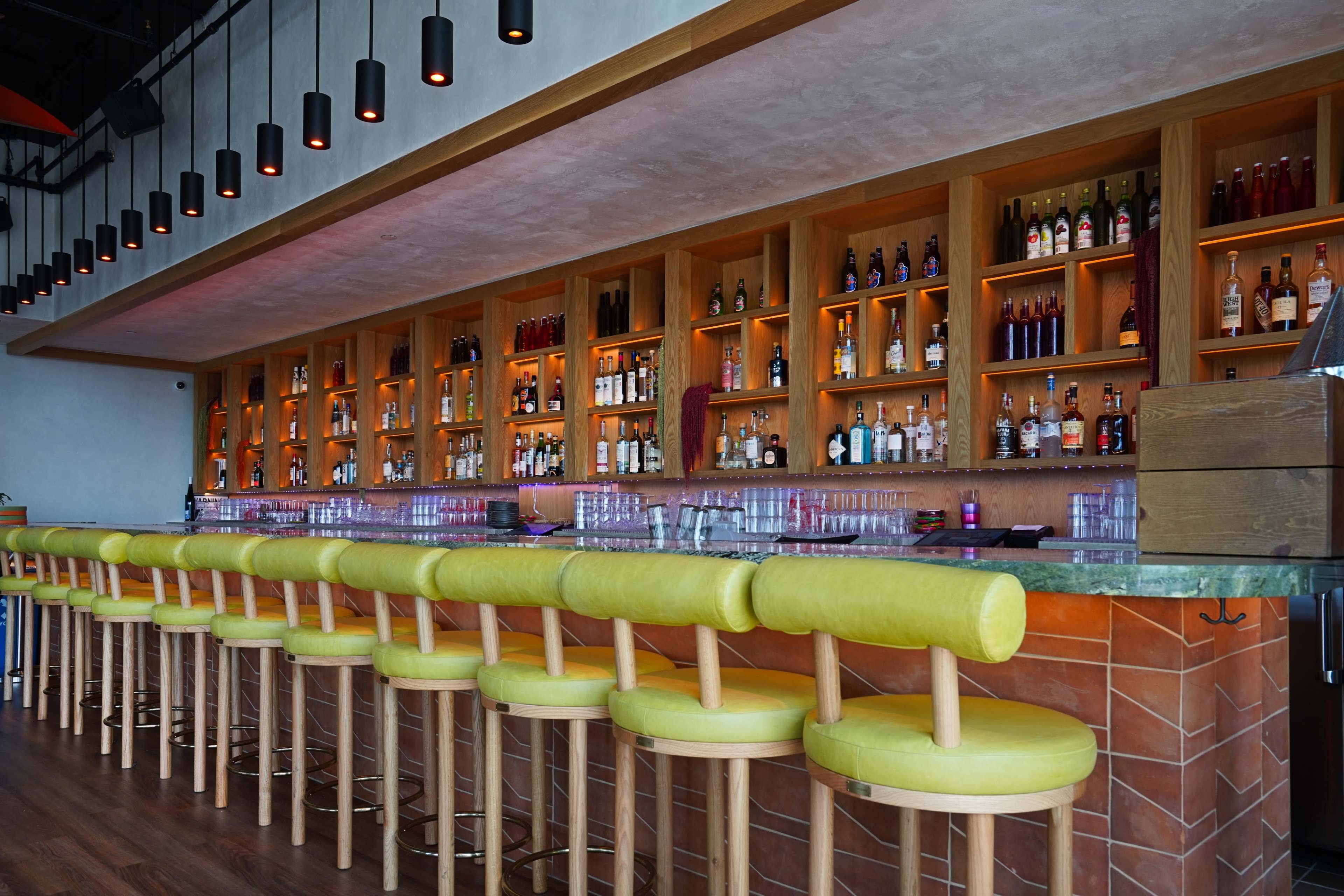 The image shows a modern bar with a long row of yellow upholstered stools in front of a wooden shelving unit filled with various bottles and illuminated by soft lighting.