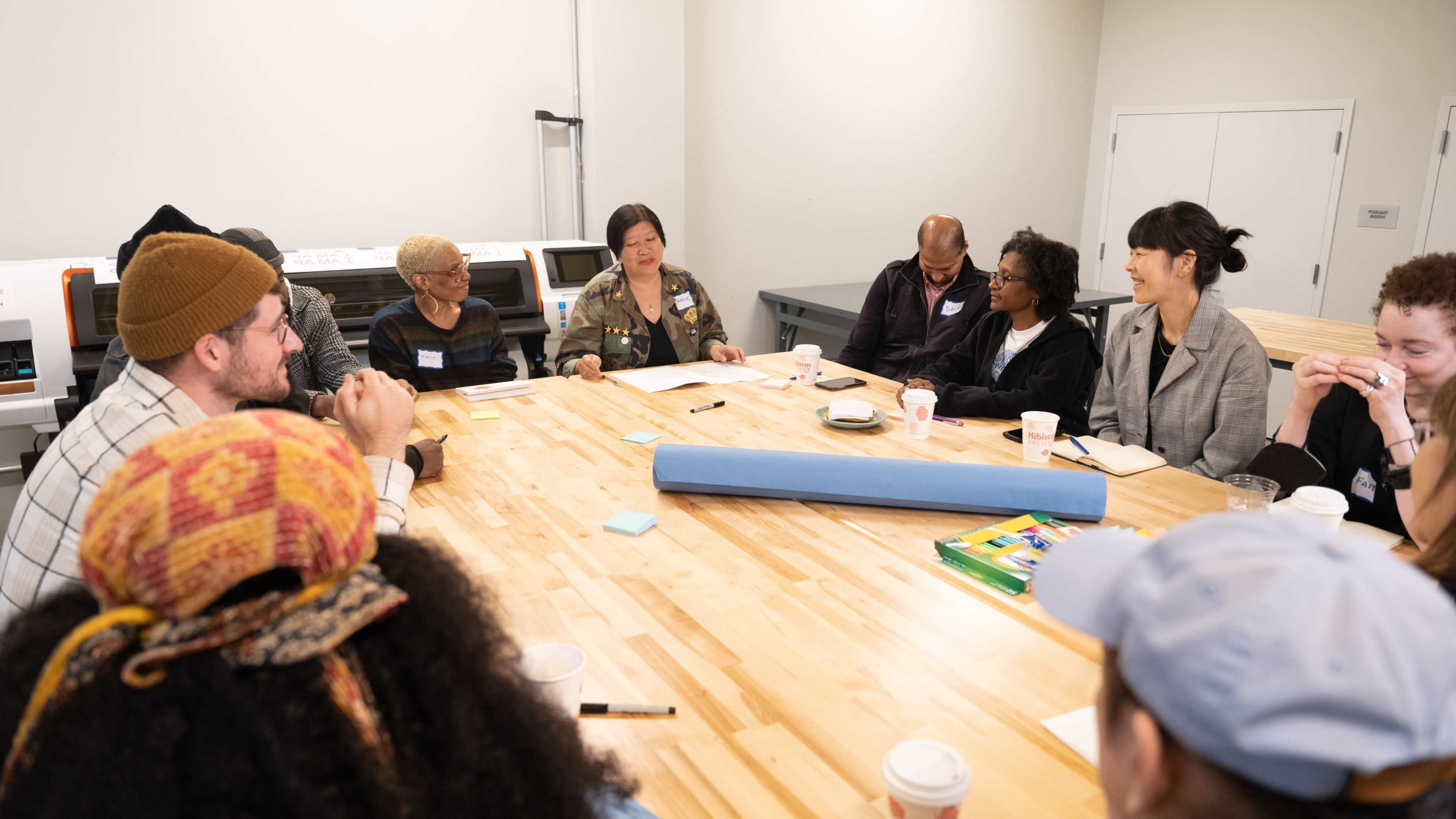 A diverse group of people sits around a large wooden table, engaged in discussion and collaborating with various materials in front of them.