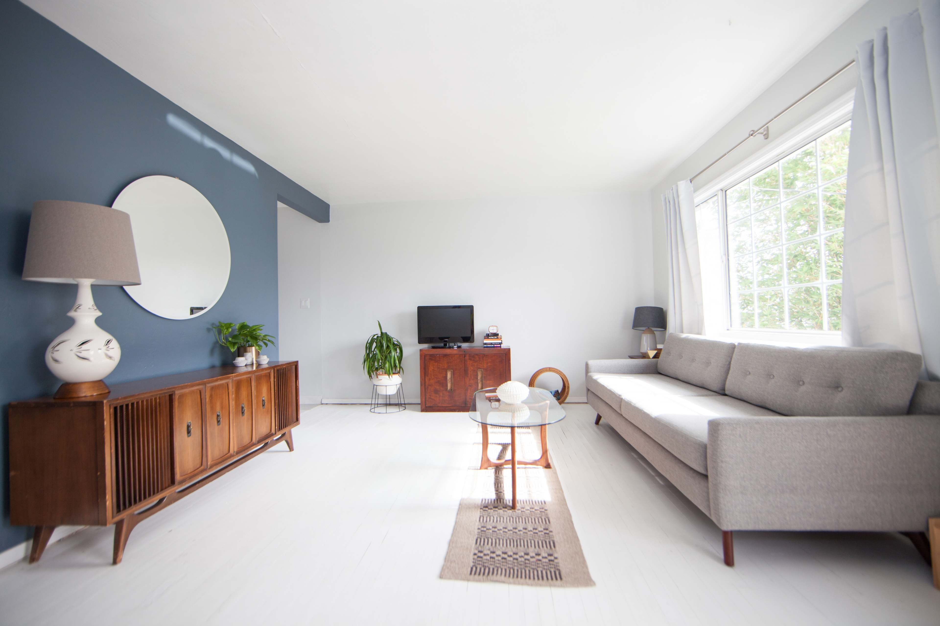A modern living room with a gray sofa, a round mirror on a blue wall, a wooden sideboard, and a coffee table positioned in front of a window.