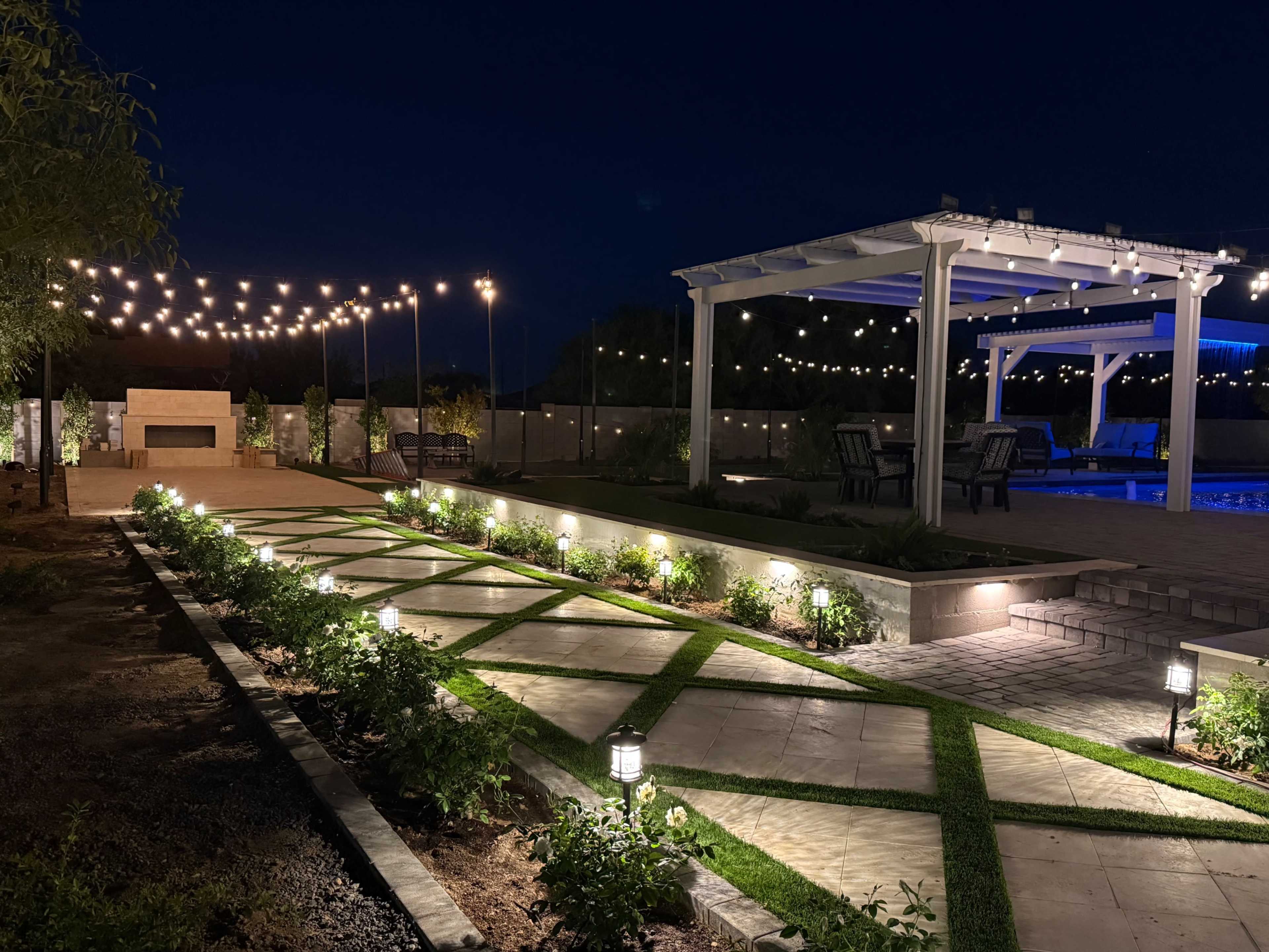 The image shows a well-lit outdoor patio area at night, featuring a pool, seating under a pergola, string lights overhead, and landscaped pathways with lights and plants.