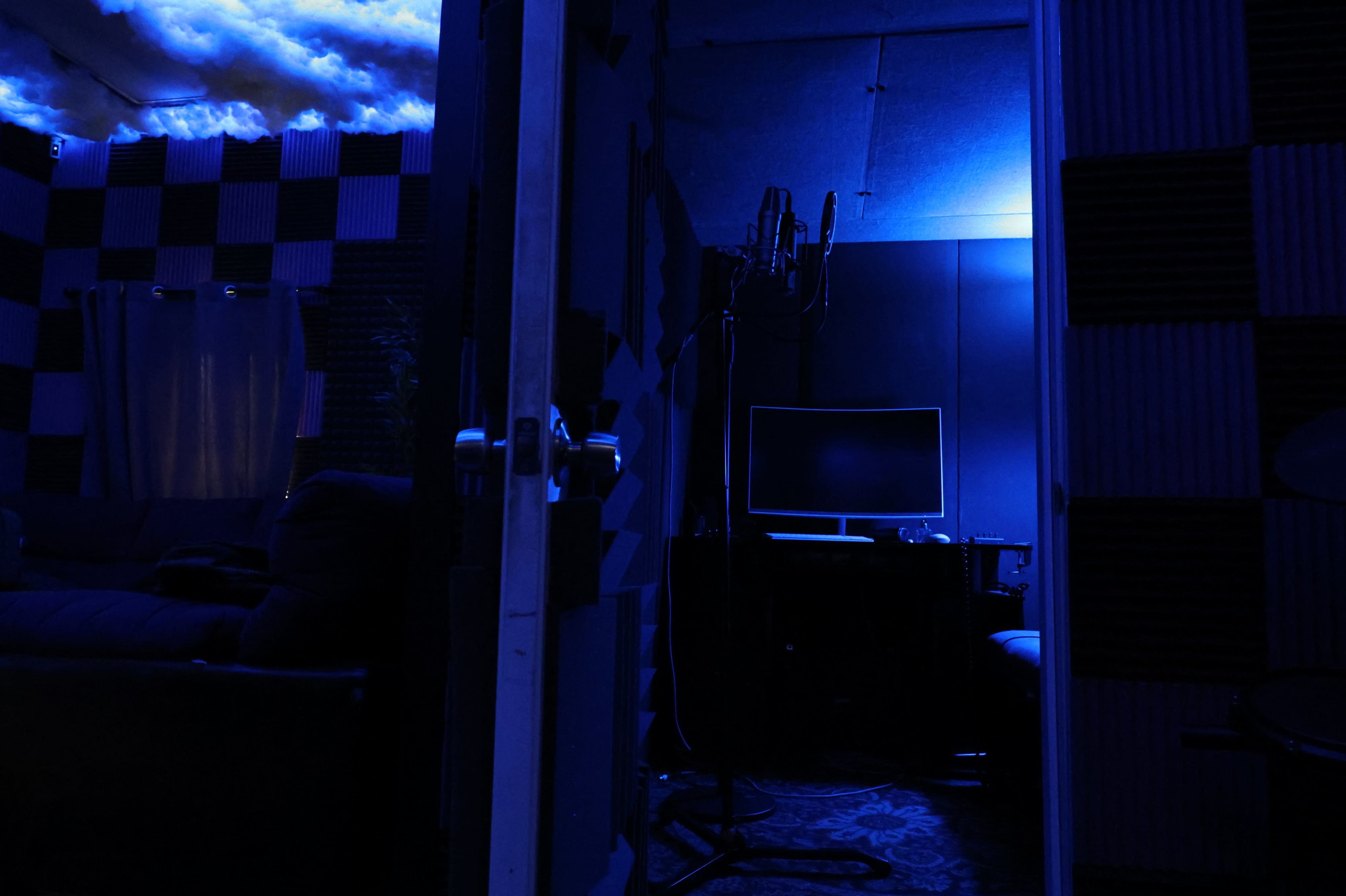 A dimly lit room with blue lighting features a microphone stand, a computer monitor, and soundproofing panels.
