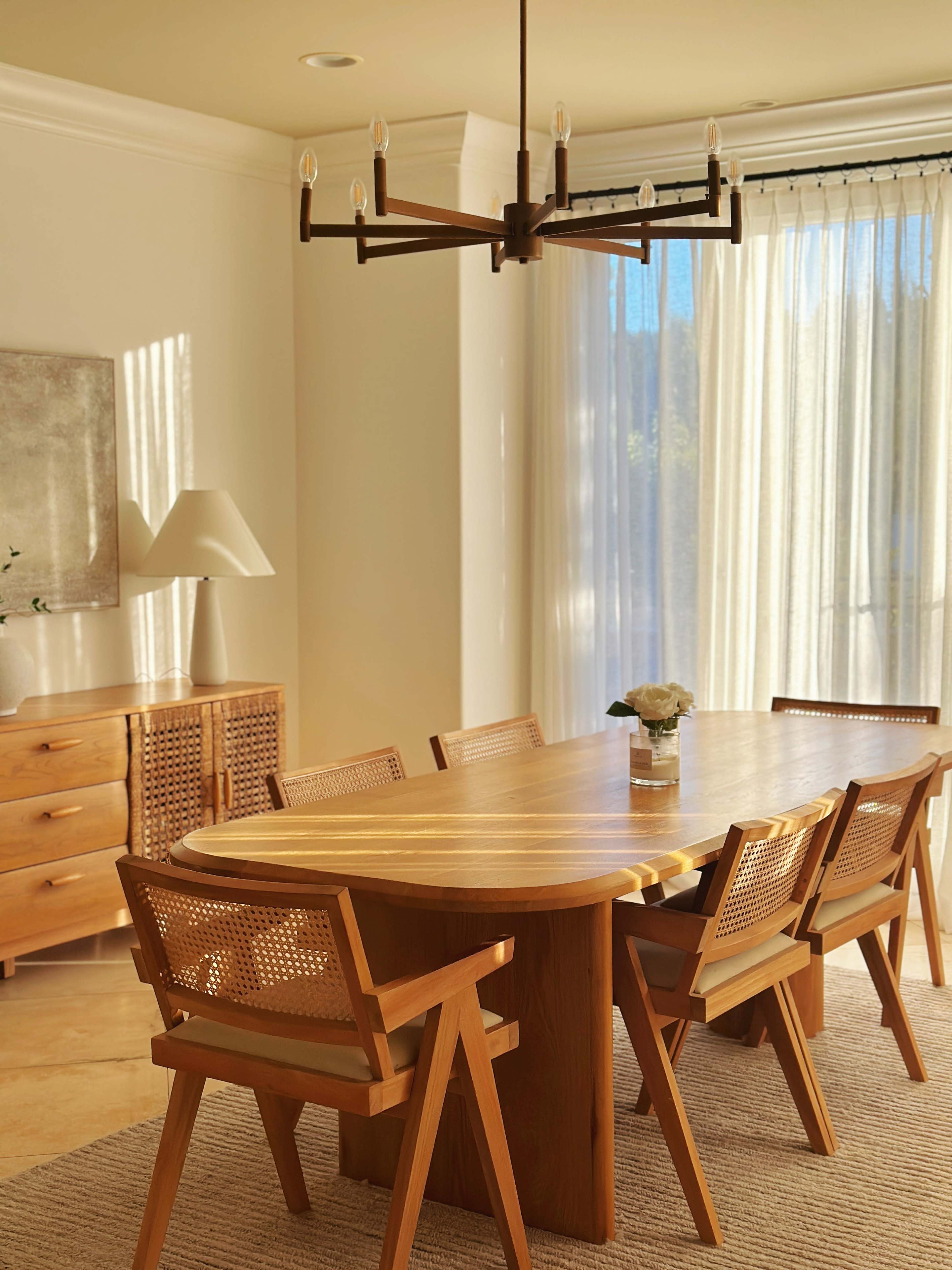 A wooden dining table with five chairs is positioned in a well-lit room featuring a chandelier and a sideboard.