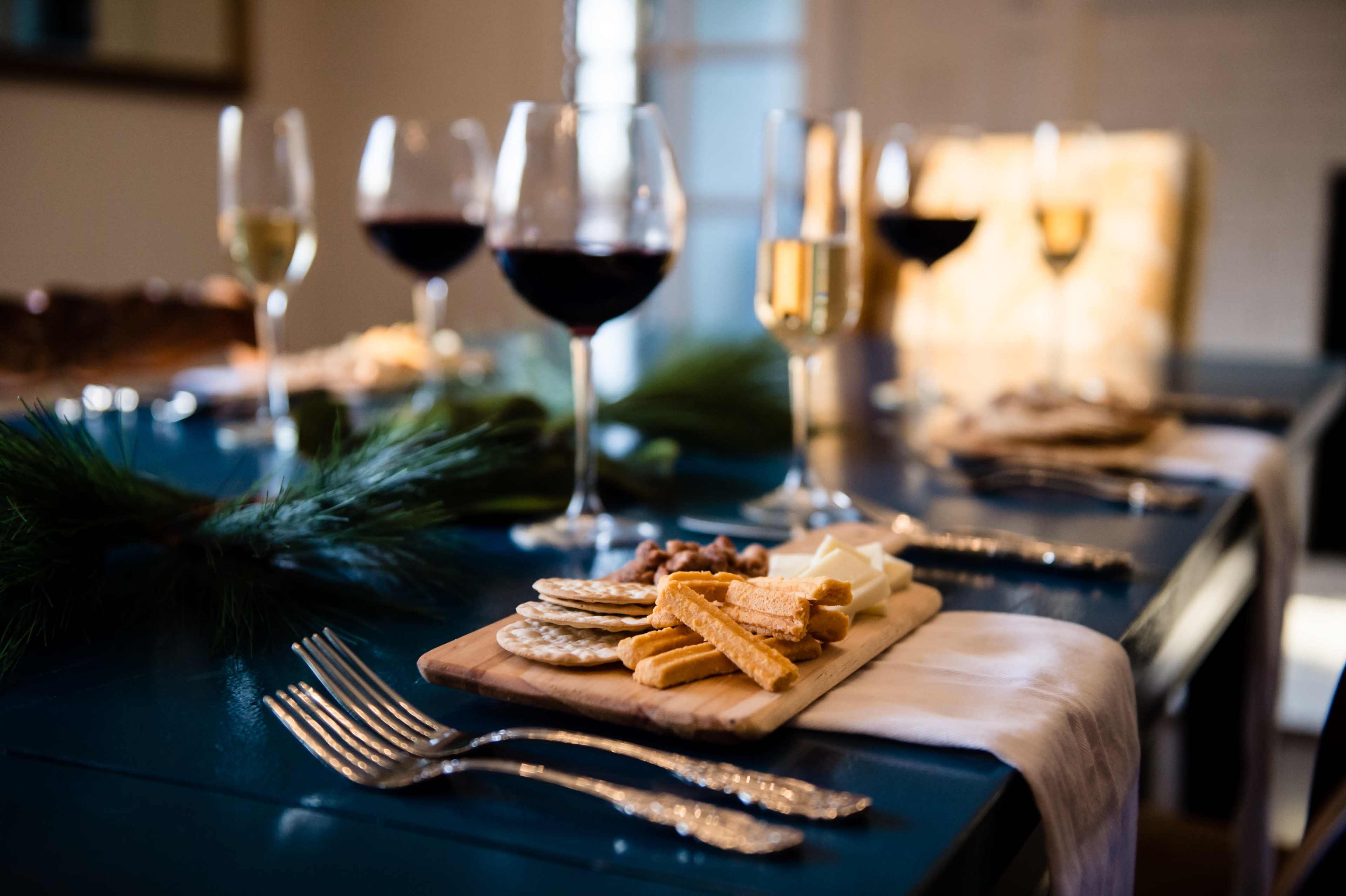 A wooden platter with an assortment of cheeses and crackers is set on a dining table adorned with wine glasses and greenery.