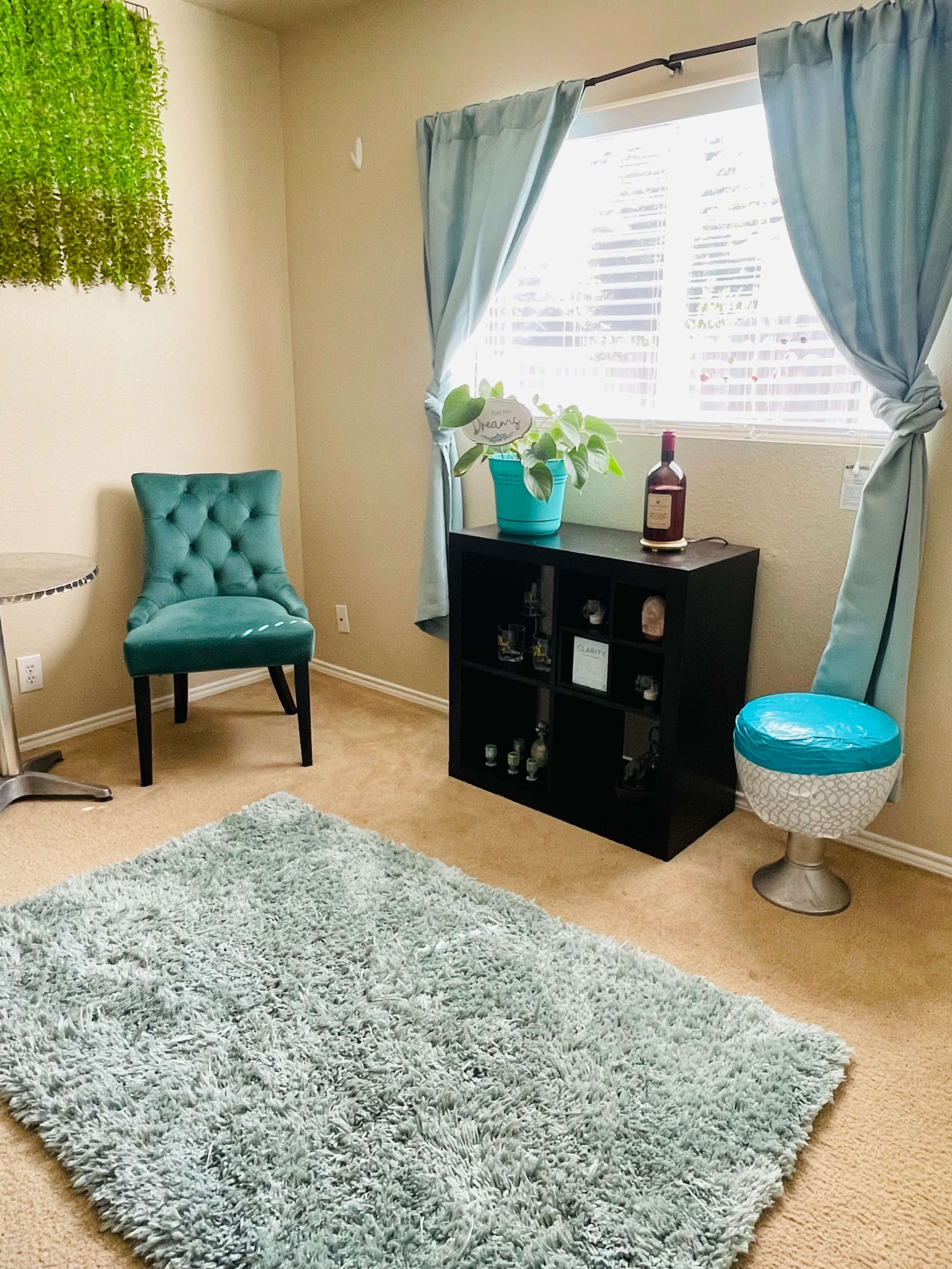 The image shows a cozy corner of a room featuring a turquoise chair, a small bookshelf with decorative items, a plant, and a round stool, all set against a light-colored wall and window with curtains.