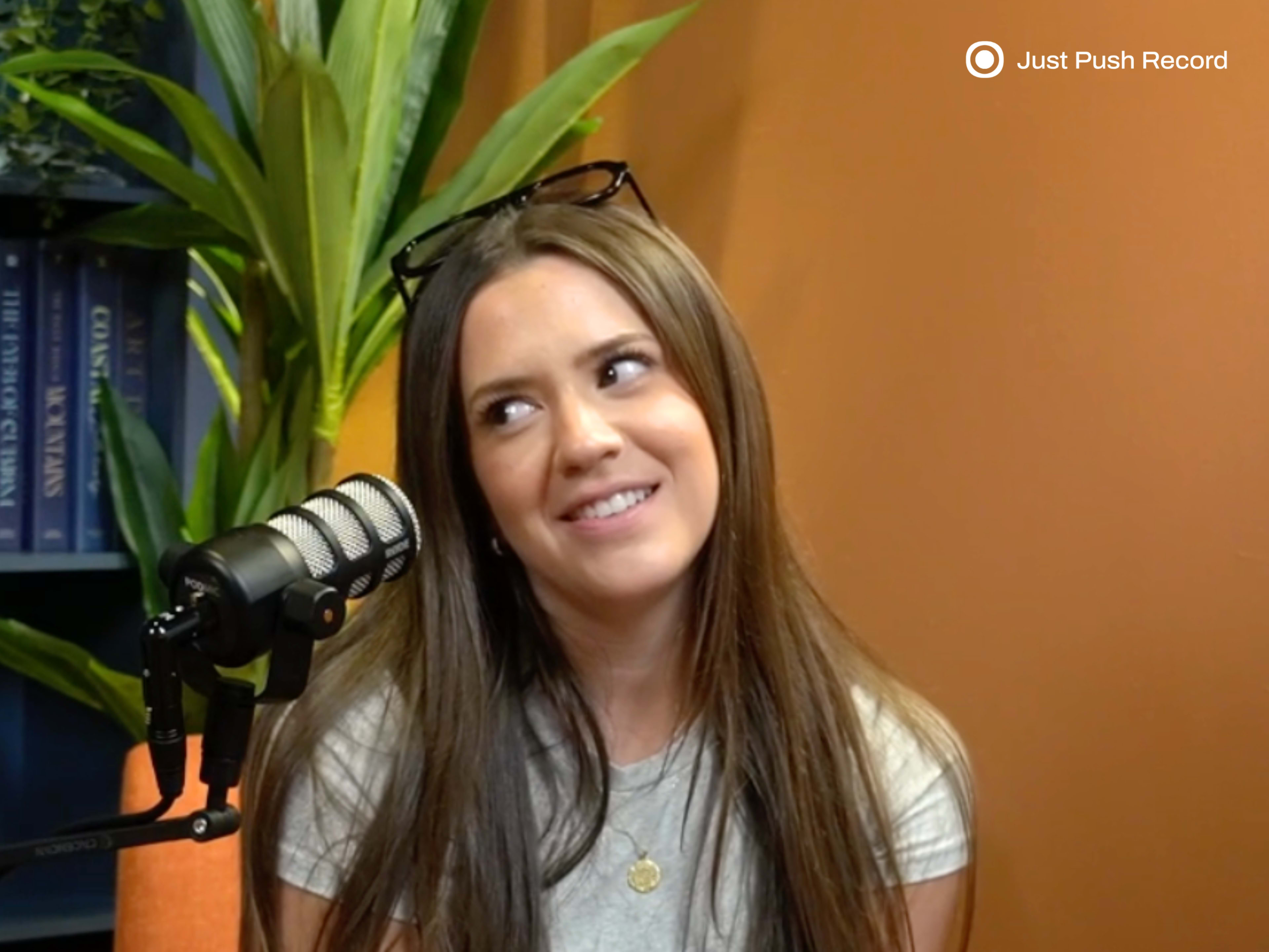 A woman with long hair sits in front of a microphone and appears to be listening or reacting during a conversation.