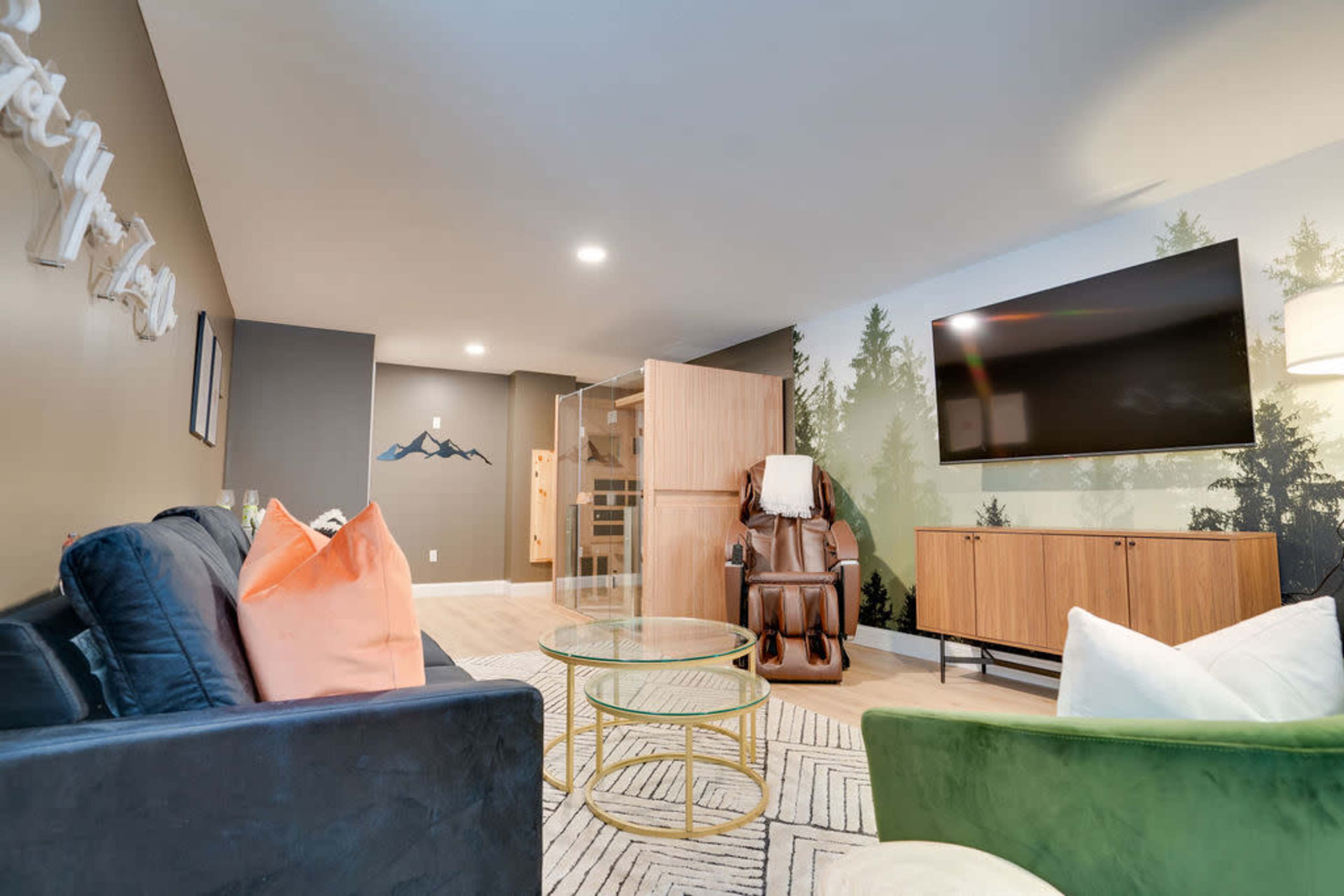 The image depicts a modern living room featuring a television mounted on the wall, a massage chair, and a wooden cabinet, with a backdrop of trees painted on the wall.