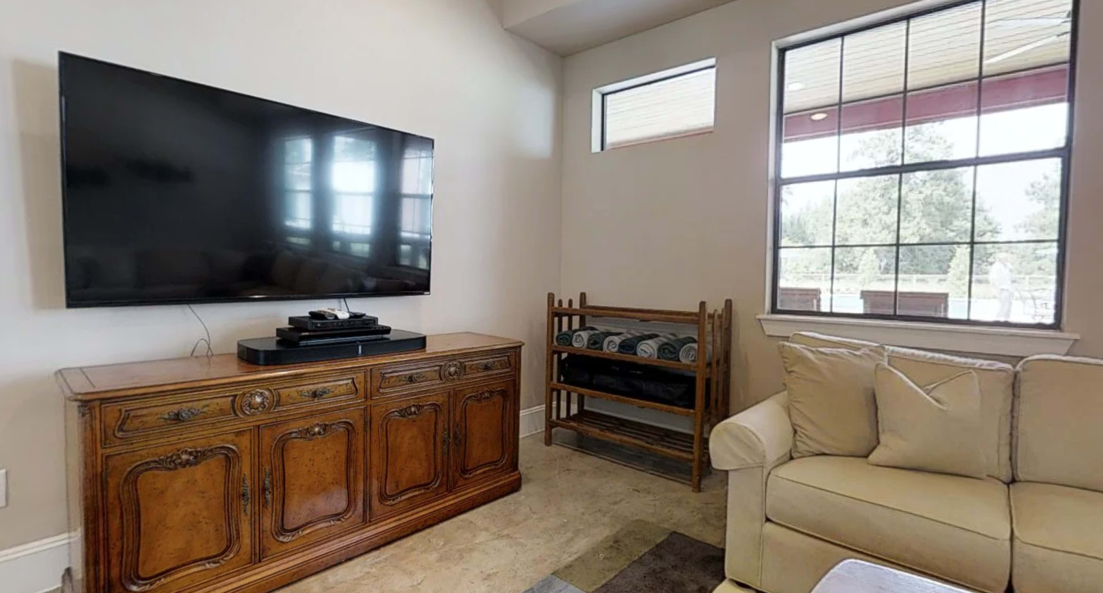 A living room featuring a large flat-screen television mounted on the wall above a wooden cabinet, with a sofa and a shelf displaying rolled-up towels beside a window.