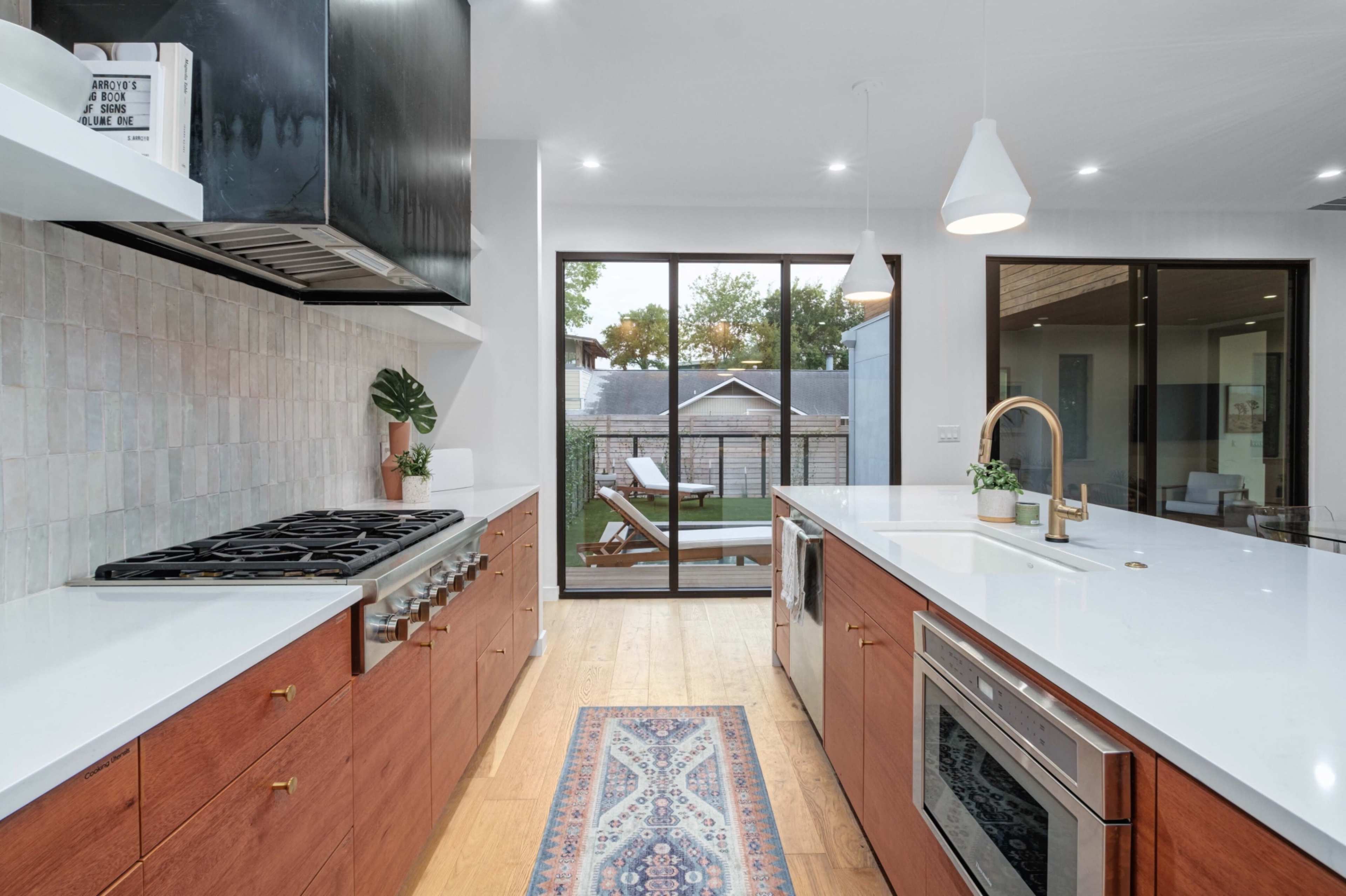 A modern kitchen features wooden cabinets, a large island with a countertop, a stainless steel gas stove, and an adjacent deck visible through large glass doors.