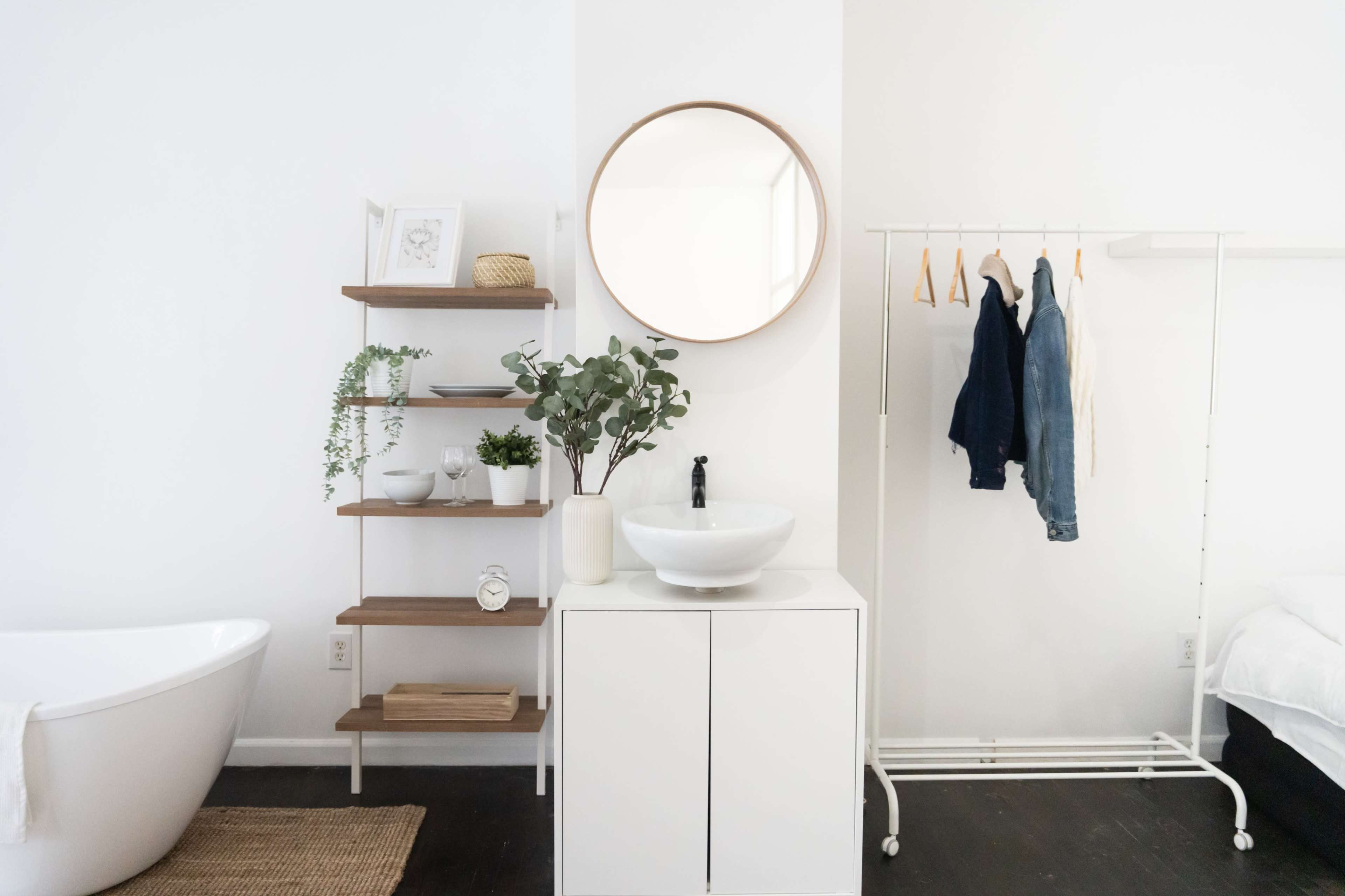 A modern bathroom space featuring a round mirror above a white sink on a minimalist cabinet, alongside a wooden shelf with decorative items and plants, and a clothing rack displaying garments.