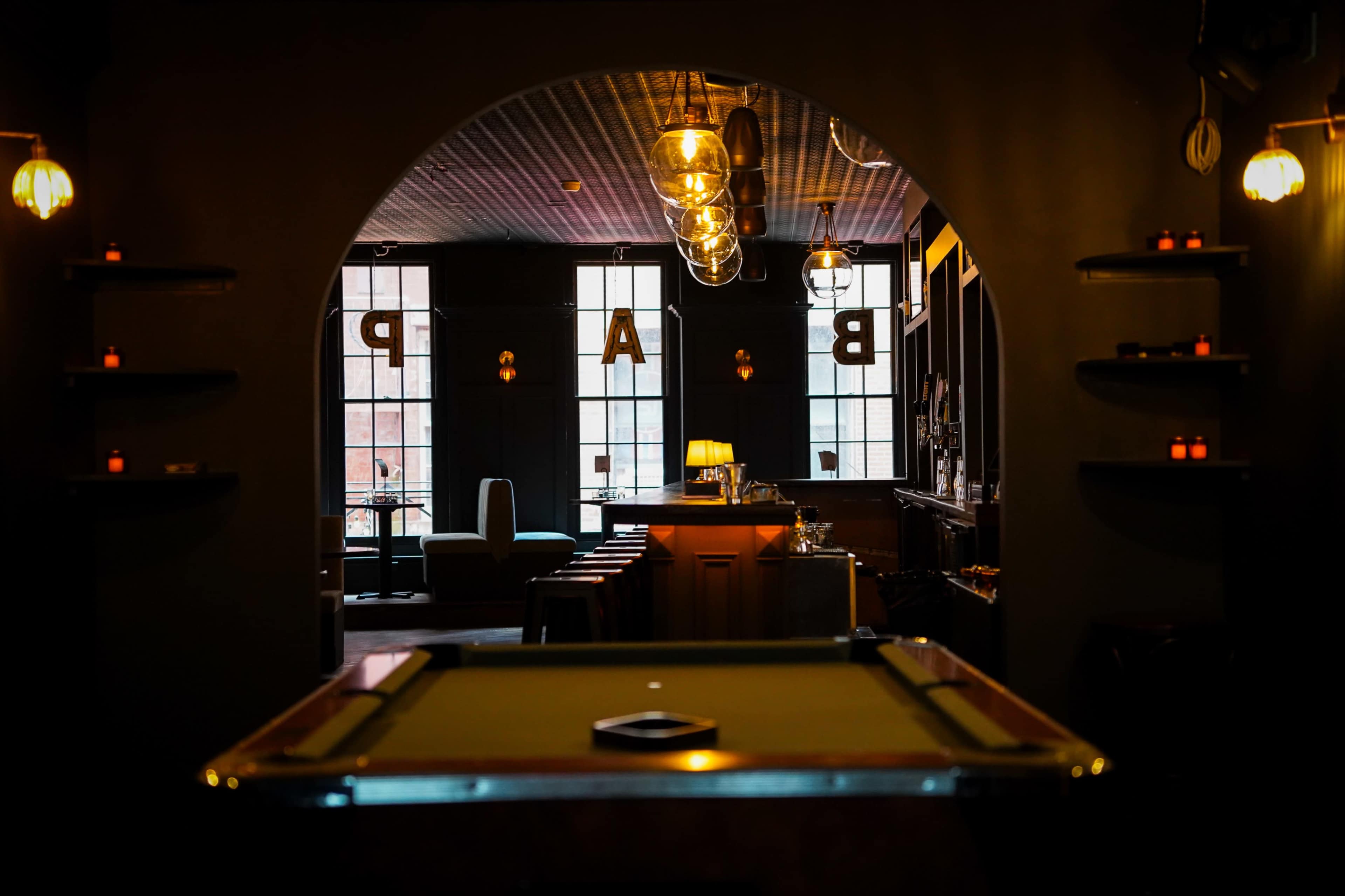 The image shows a dimly lit bar setting viewed through an archway, with a pool table in the foreground and shelves holding candles along the walls.