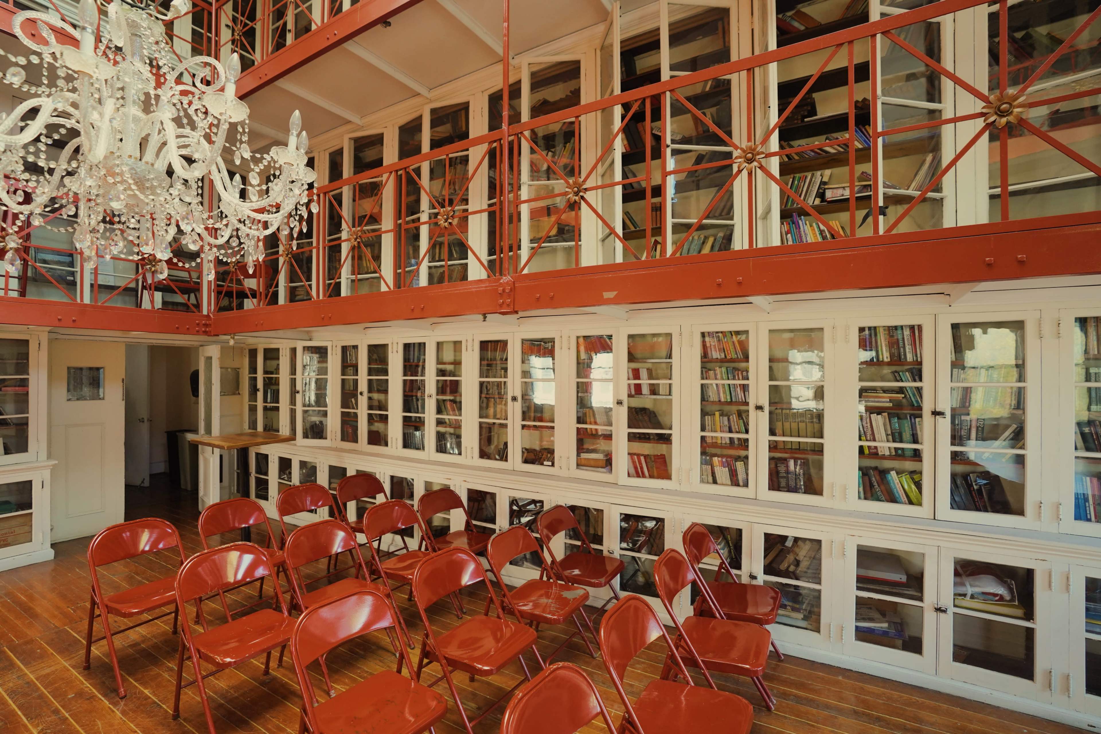 A bright, open library space featuring tall shelves filled with books and a set of red chairs arranged in front.