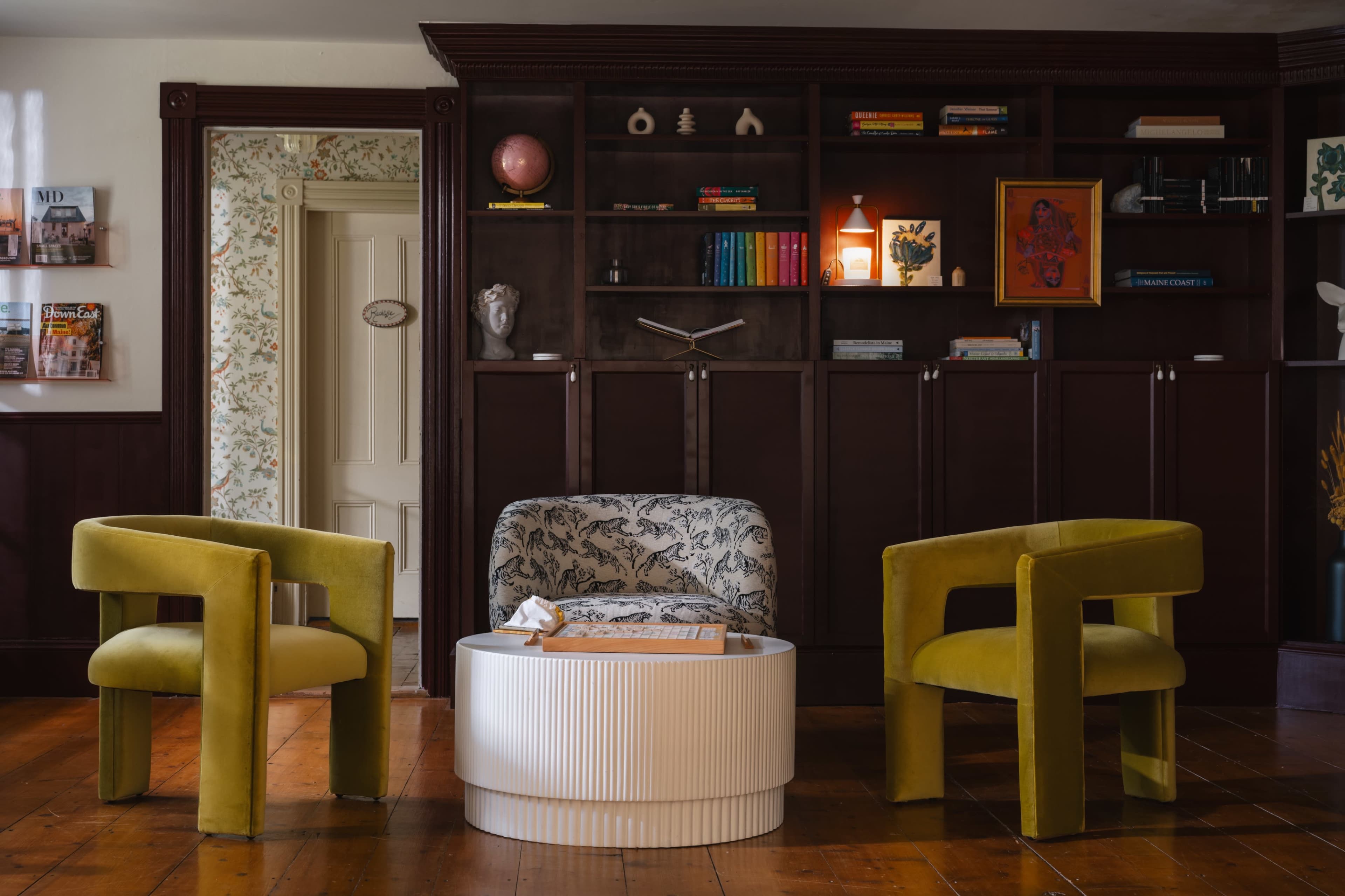 The image features a cozy living room setup with two modern chairs in yellow and green flanking a white round coffee table, against a backdrop of a dark wooden shelf filled with decorative items and books.