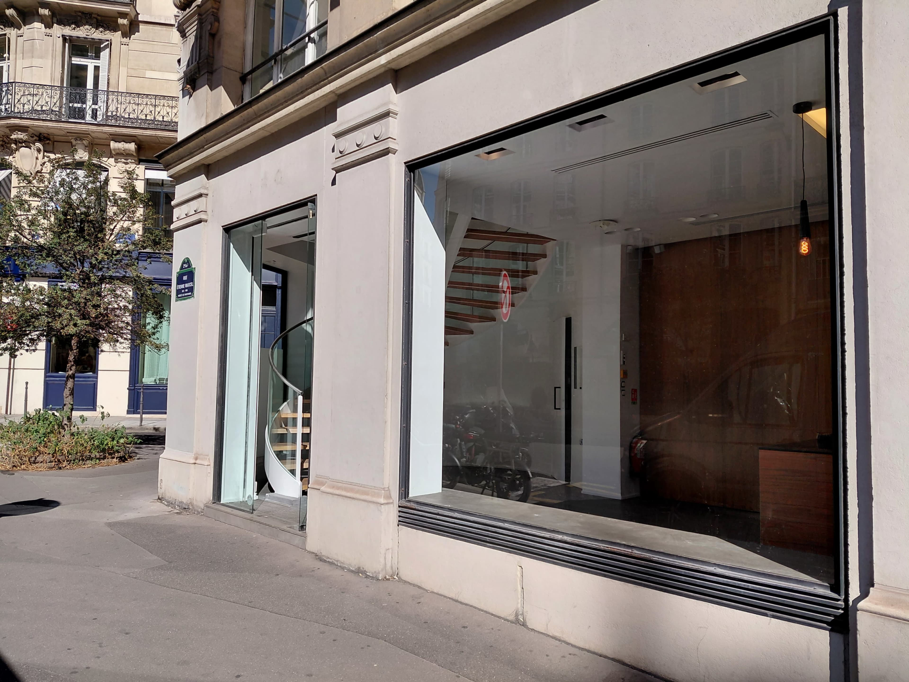 The image shows a modern retail space with large glass windows, a wooden interior staircase, and a minimalist design located on a street corner.
