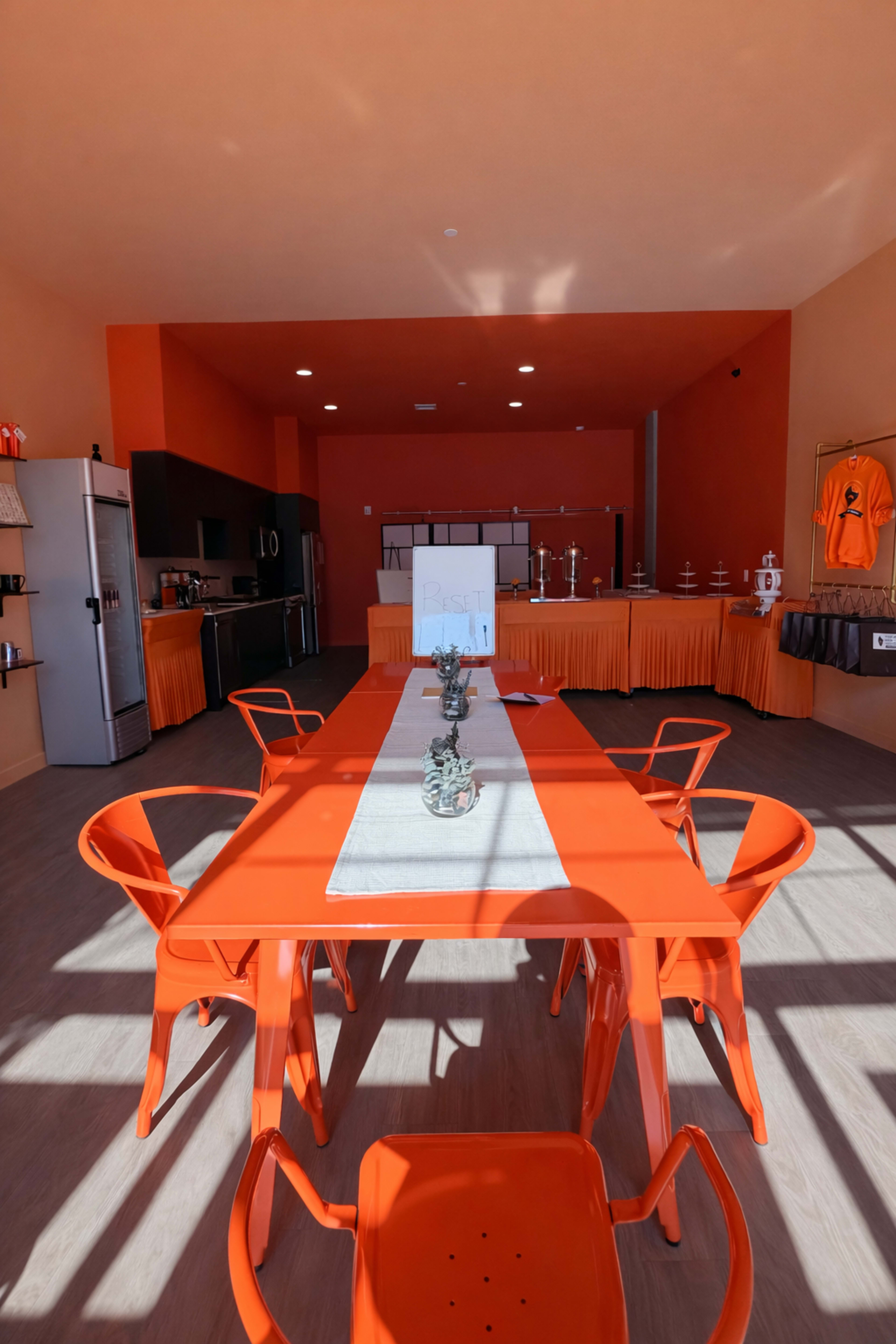 The image shows a brightly colored room with an orange theme, featuring a long table surrounded by chairs, and a kitchen area in the background.
