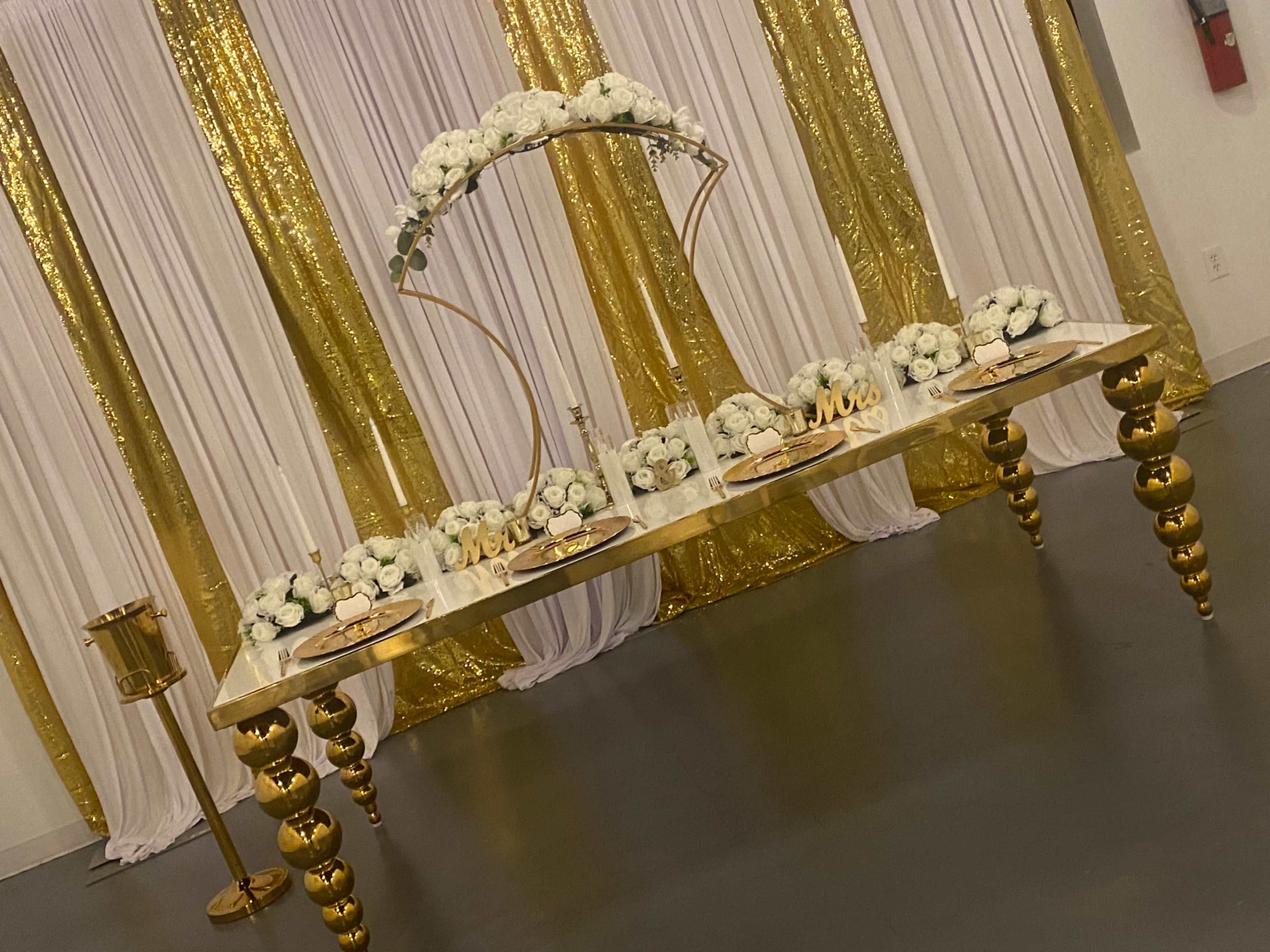 The image shows a decorated table with a gold arch and arrangements of white flowers, set against a backdrop of golden and white drapes.