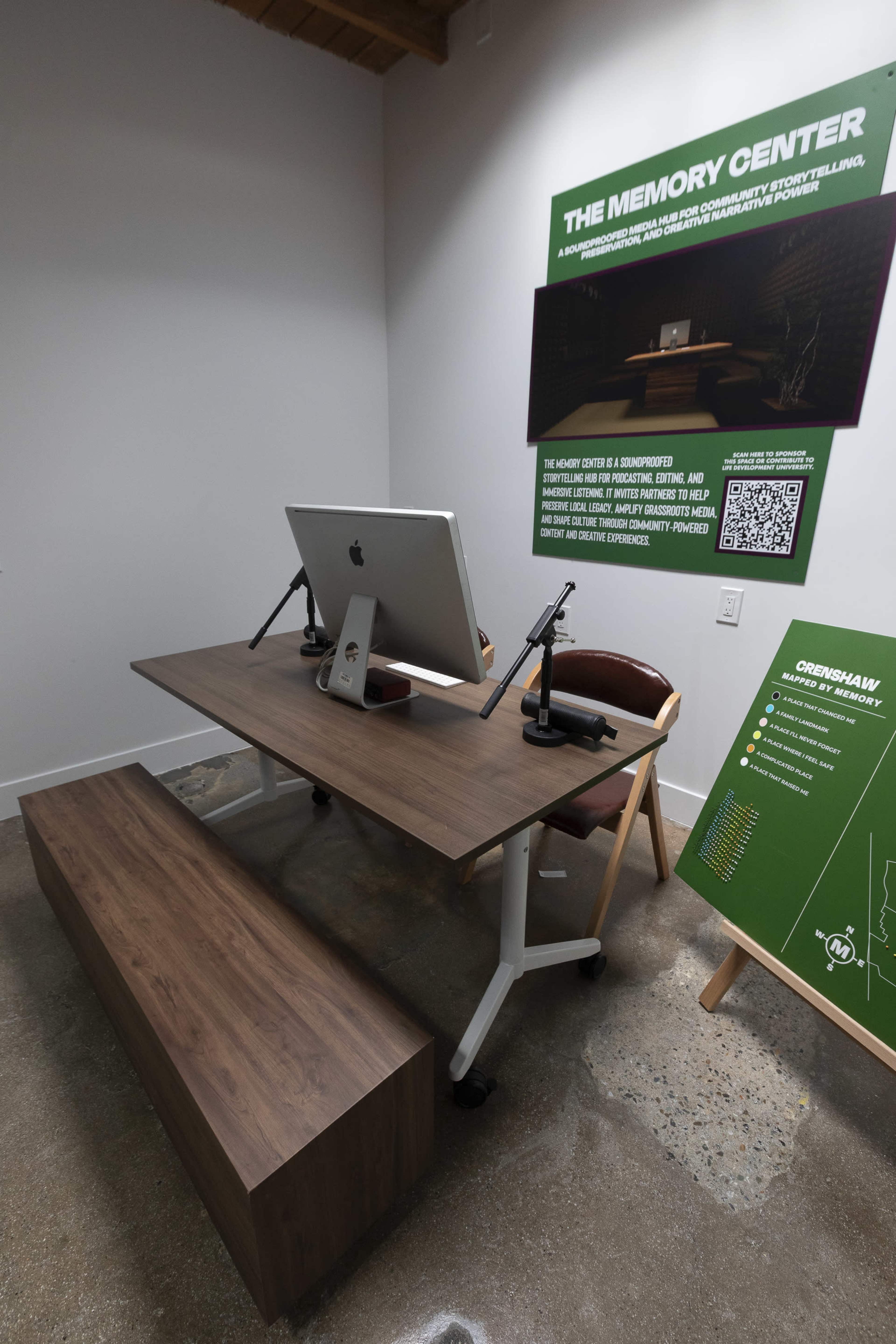 The image shows a modern workspace featuring a wooden desk with an iMac on it, accompanied by two microphones and a long wooden bench for seating.