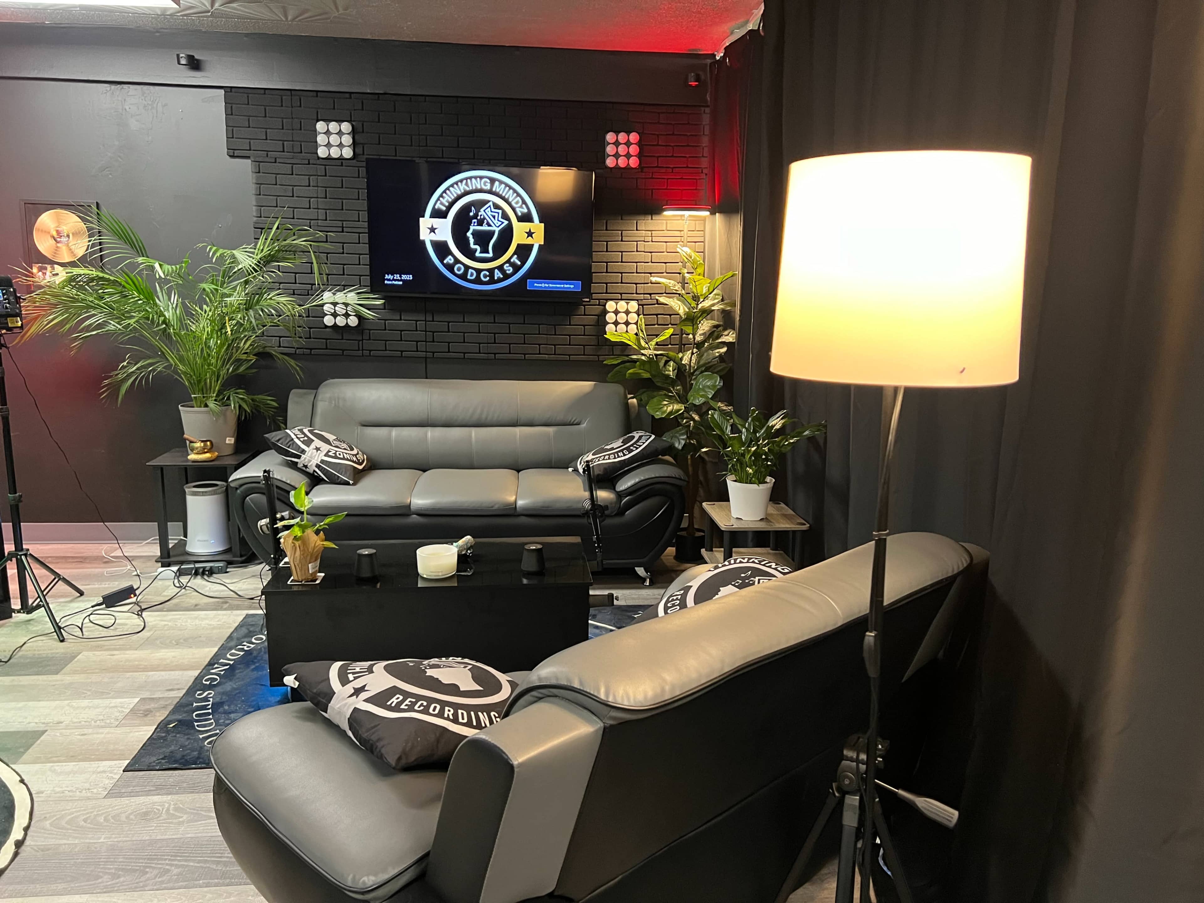 A cozy recording studio setup with two gray sofas facing a coffee table, surrounded by plants and featuring a black backdrop with a television display.