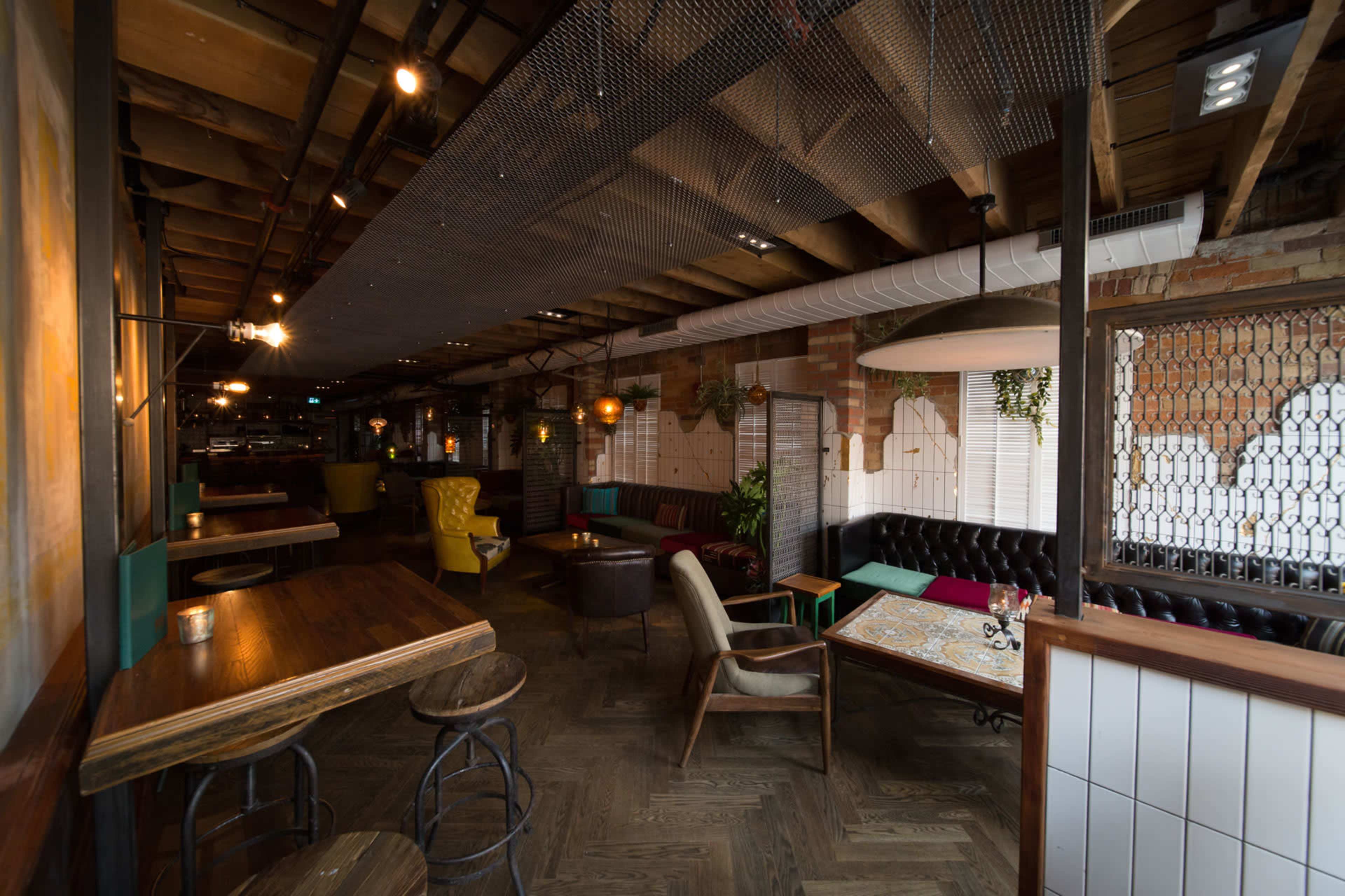 The image shows a stylish restaurant interior with a mix of seating options, wooden tables, and exposed beams overhead.