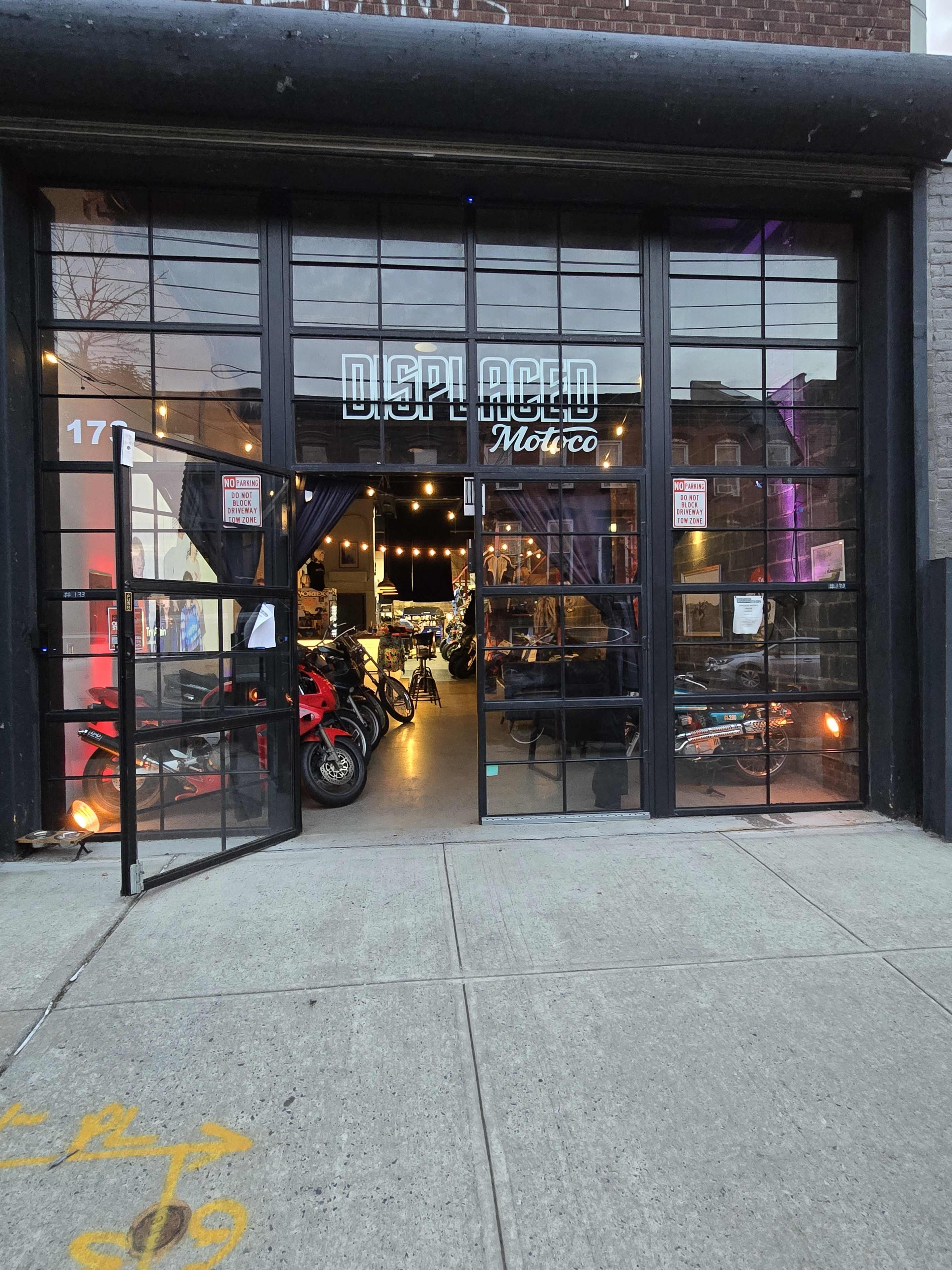The image shows the entrance of a shop called "Displaced Moto," featuring a large glass door that opens to a brightly lit interior displaying motorcycles and apparel.