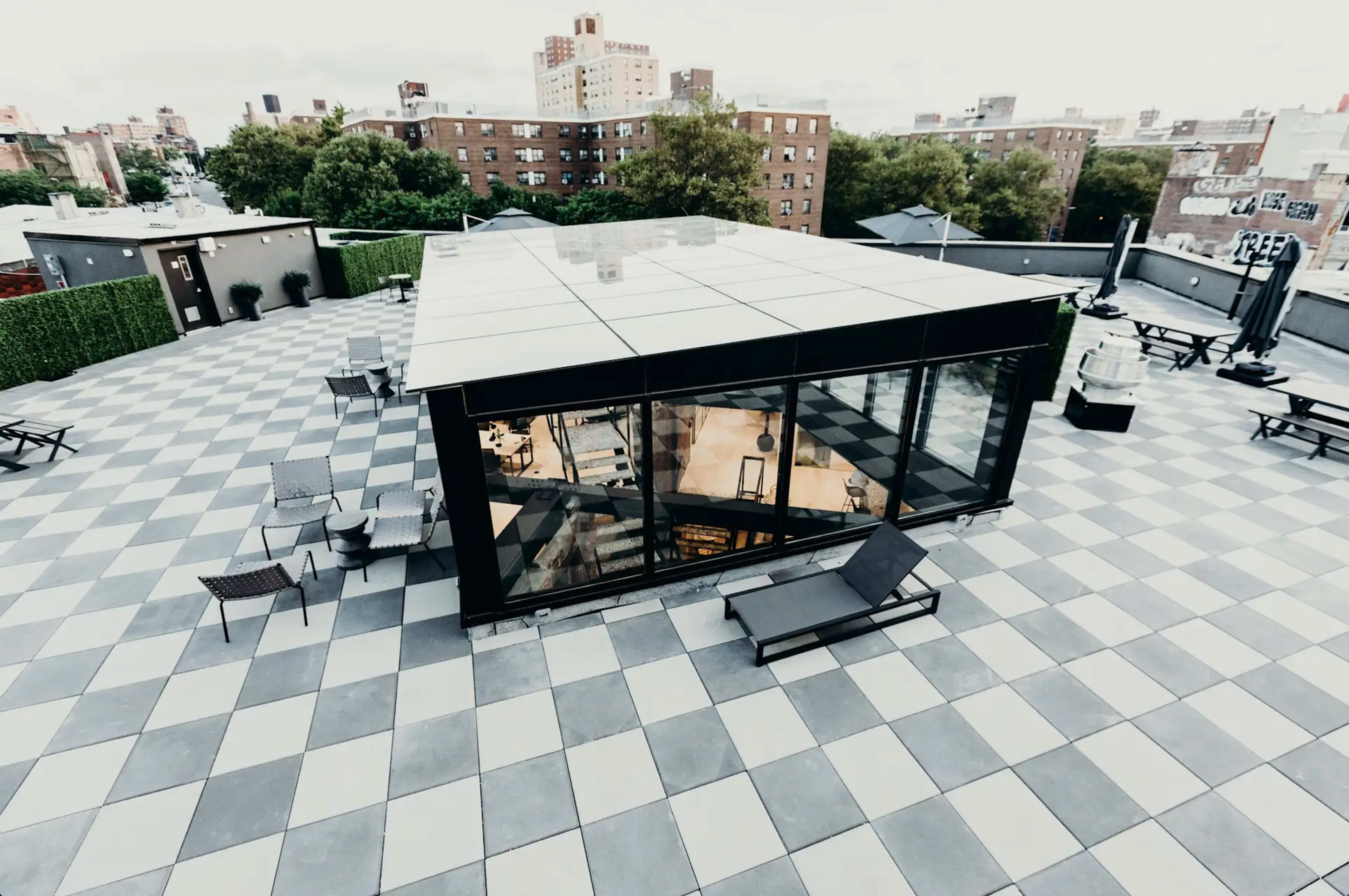 A modern glass pavilion sits atop a rooftop covered in gray and white tiled squares, surrounded by seating areas and city buildings in the background.