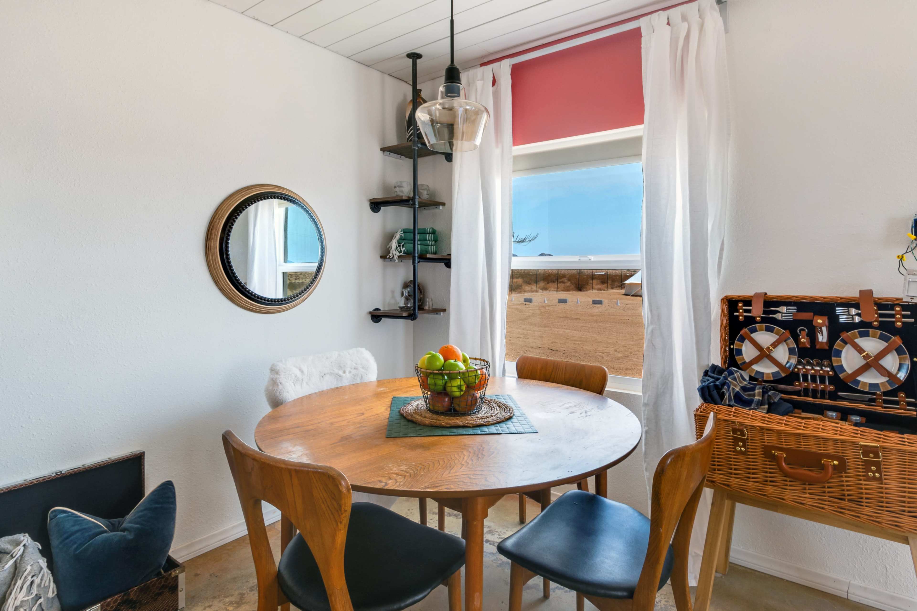 A round wooden table with four chairs is set in a bright room featuring a window, a mirror, and a wicker basket.